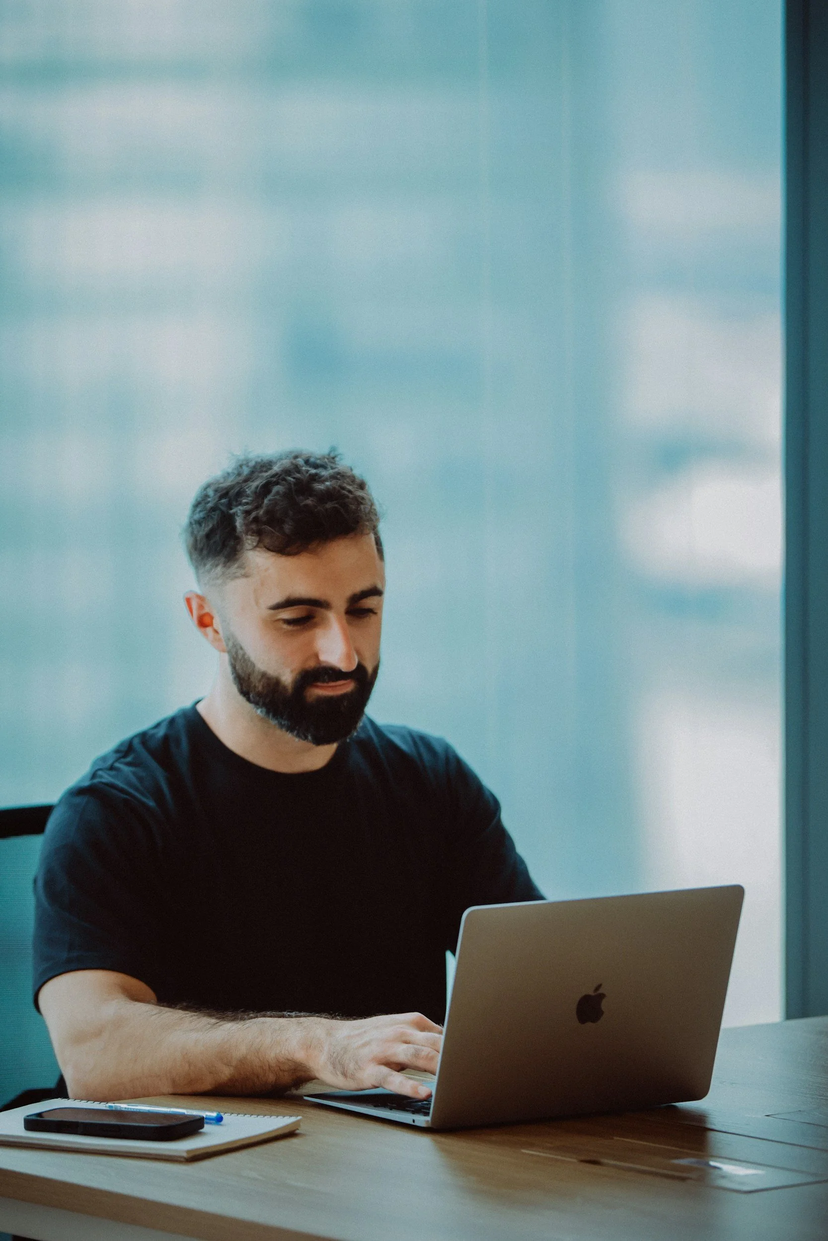 A man with dark hair and a beard sits at a table on a laptop. He represents someone who is looking for therapy for addiction in New York or Pennsylvania.