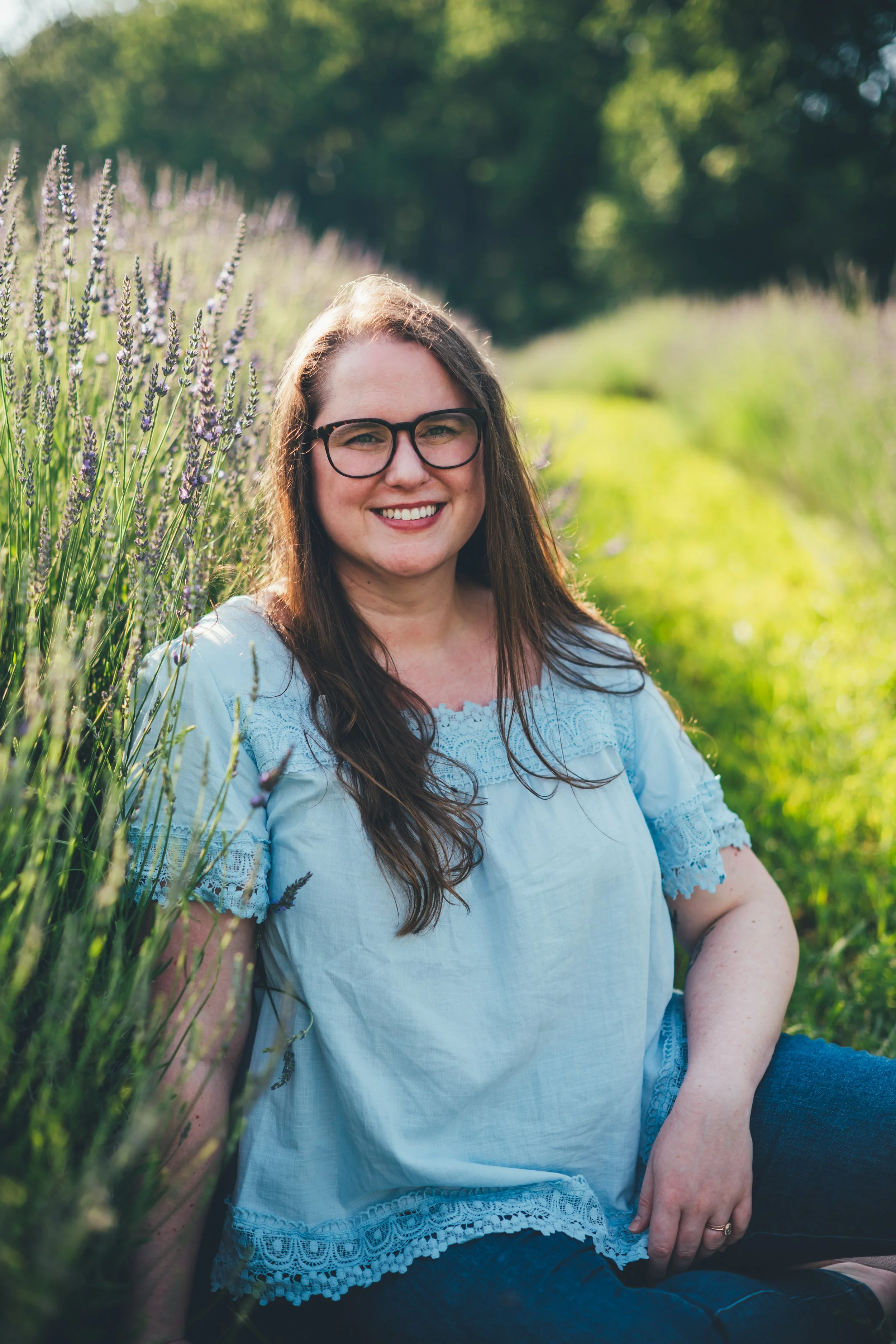 A woman with brown hair, glasses, and a light blue shirt sits smiling in a lavender field and starting directly at the camera. She is a therapist for high achieving professionals with anxiety, burnout, and problematic alcohol and other substance use.