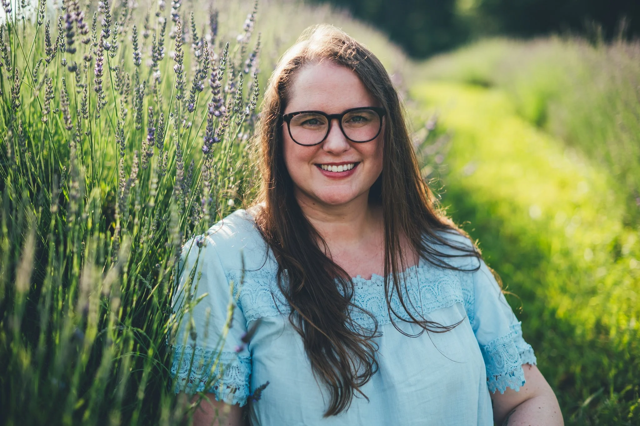 A woman sits in a lavender field and smiles at the camera. She is wearing a light blue shirt and glasses. She is a therapist who specializes in helping millennials navigate burnout and anxiety related to career and other life stressors.