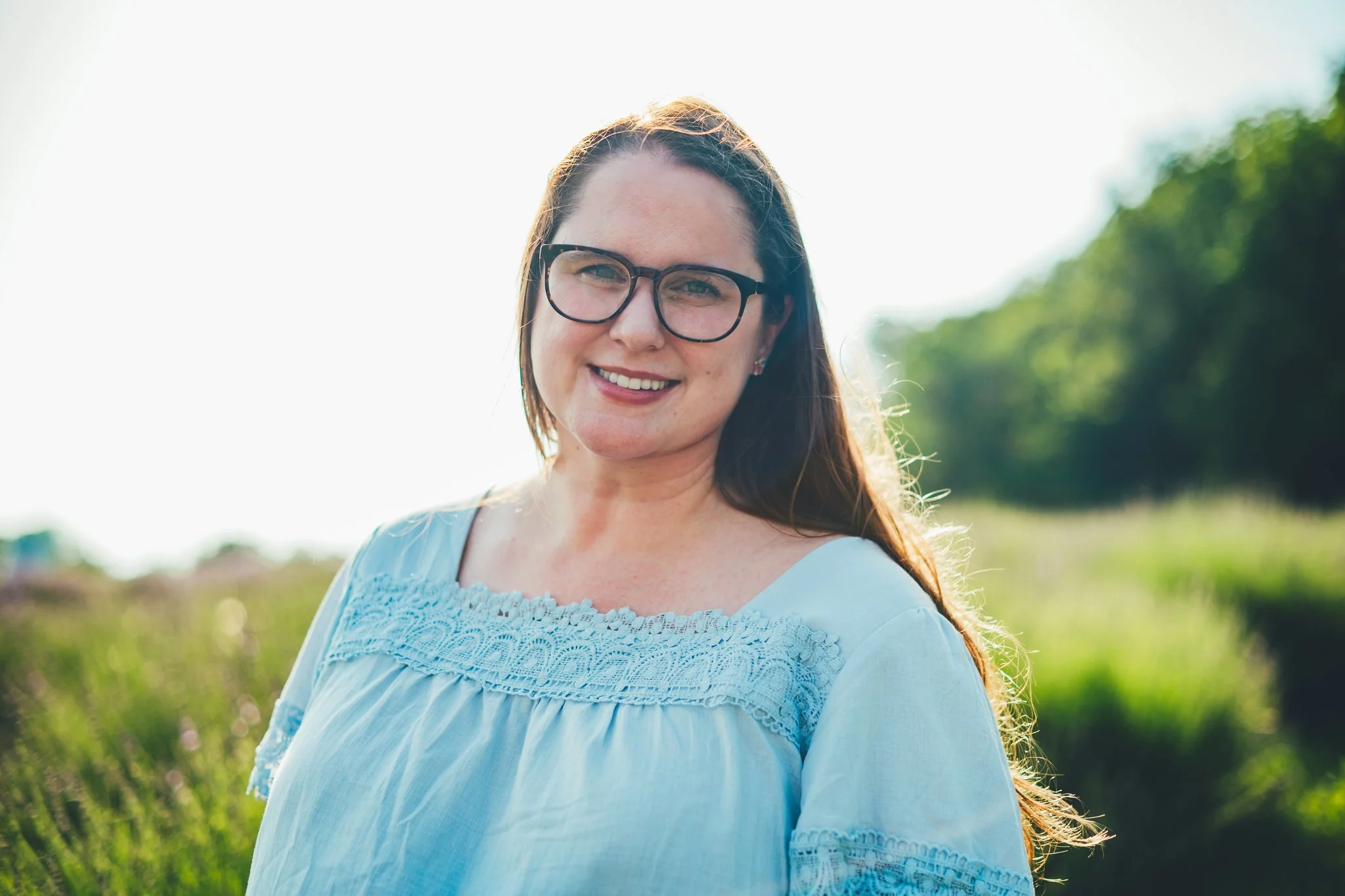 A woman wearing glasses stands in a lavender field on a sunny day. She is a therapist for burnout and anxiety and specializes working with high-achieving professionals.