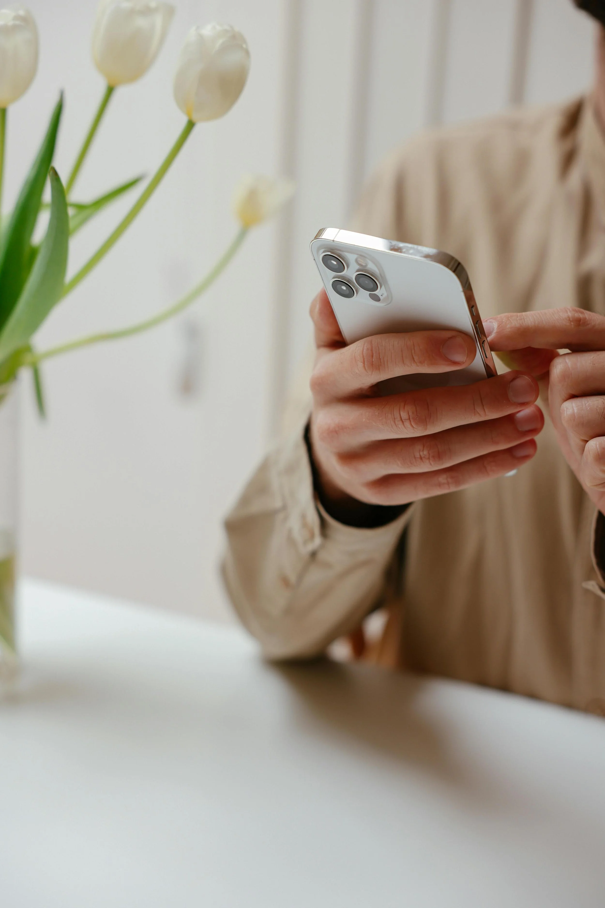 Person holding a white smartphone with triple camera setup, sitting at a white table with a vase of white tulips nearby.