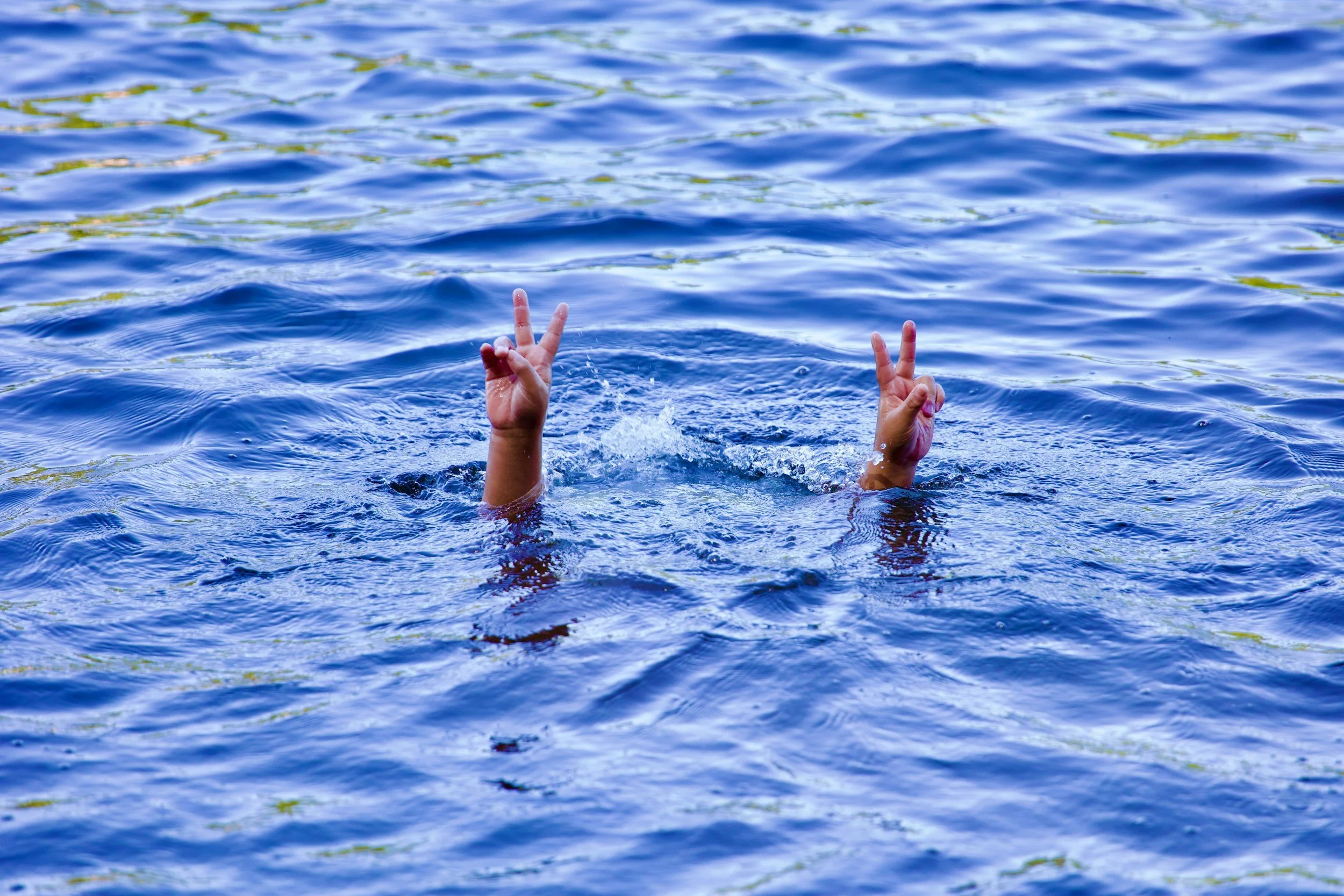 Deux mains sortent de l'eau en faisant un signe de paix dans un corps d'eau bleu.