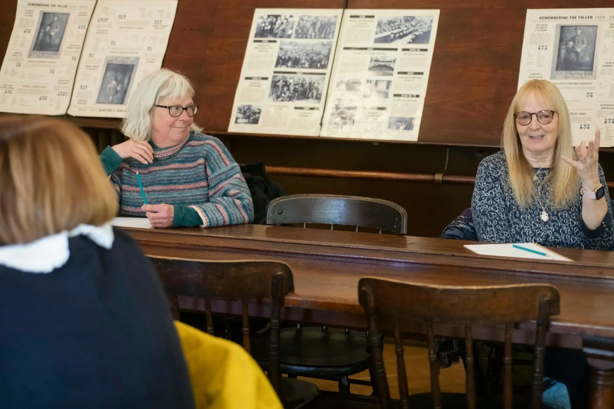 Deux femmes âgées discutent lors d'une rencontre, assises derrière une longue table en bois. Un tableau d'affichage avec des documents historiques est visible en arrière-plan.
