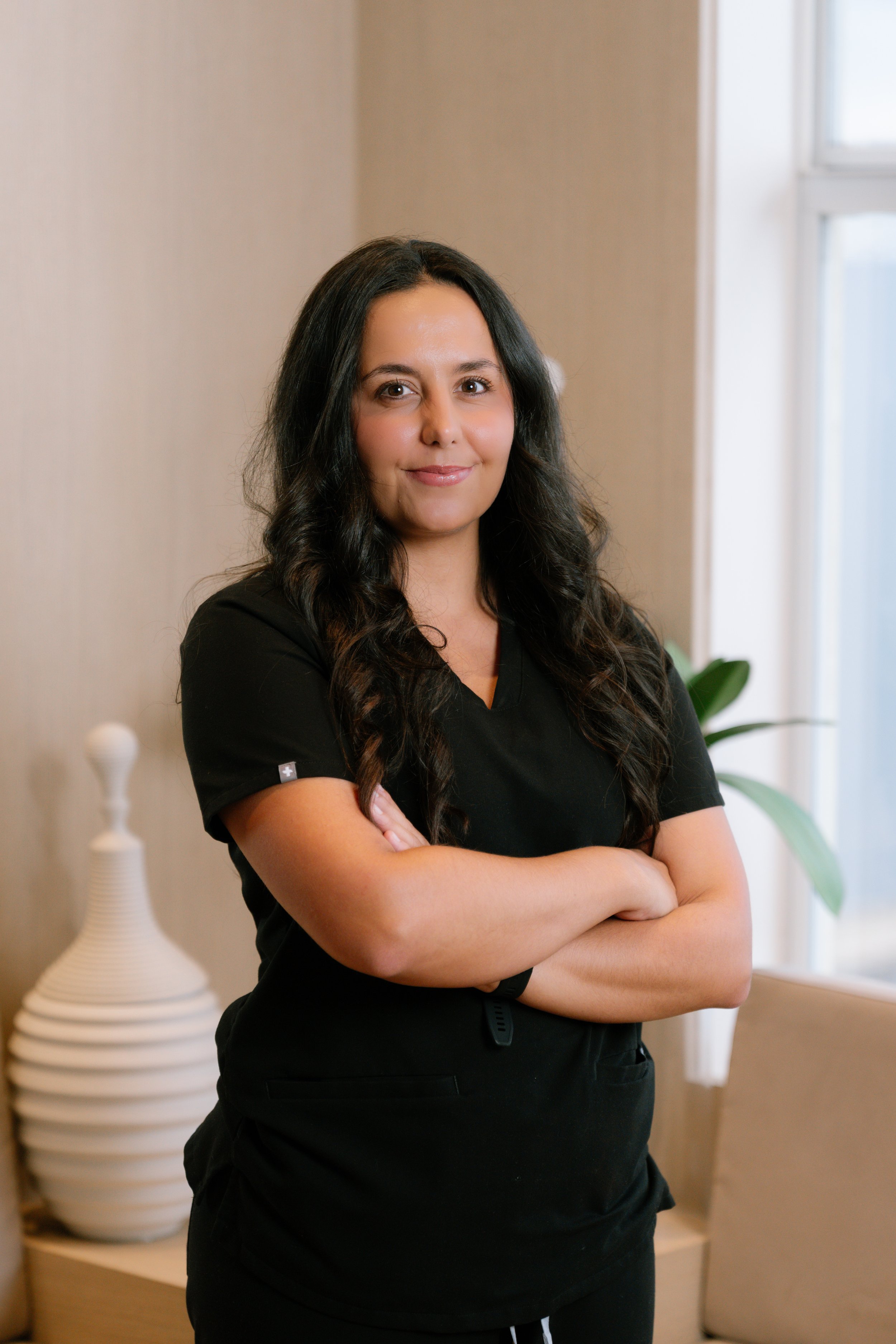 A woman in black scrubs standing with her hand on her hip in a well-lit room with large windows and a potted plant in the background.