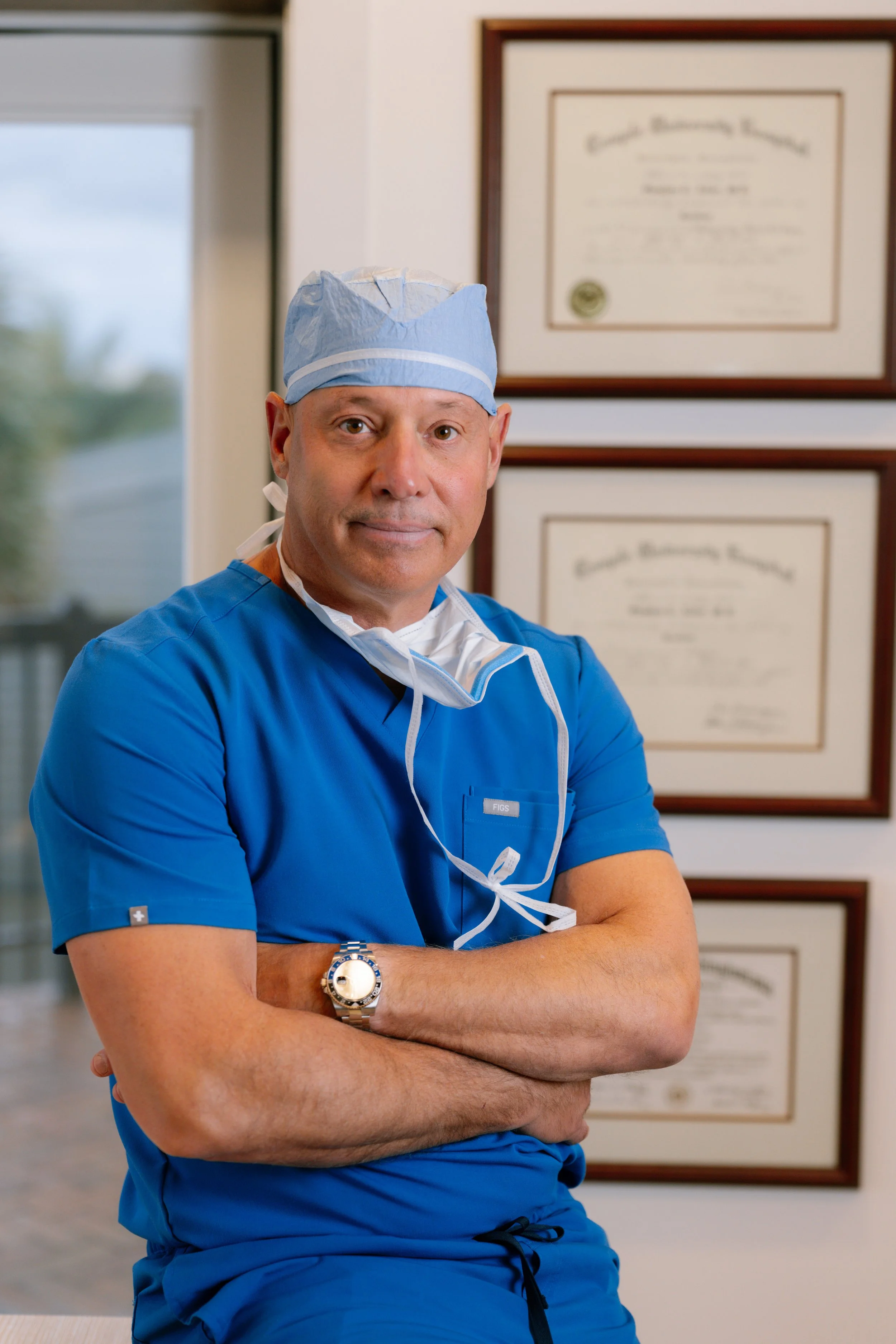 A male doctor in blue scrubs, wearing a surgical cap and a wristwatch, standing with arms crossed in a medical office with framed certificates on the wall behind him.
