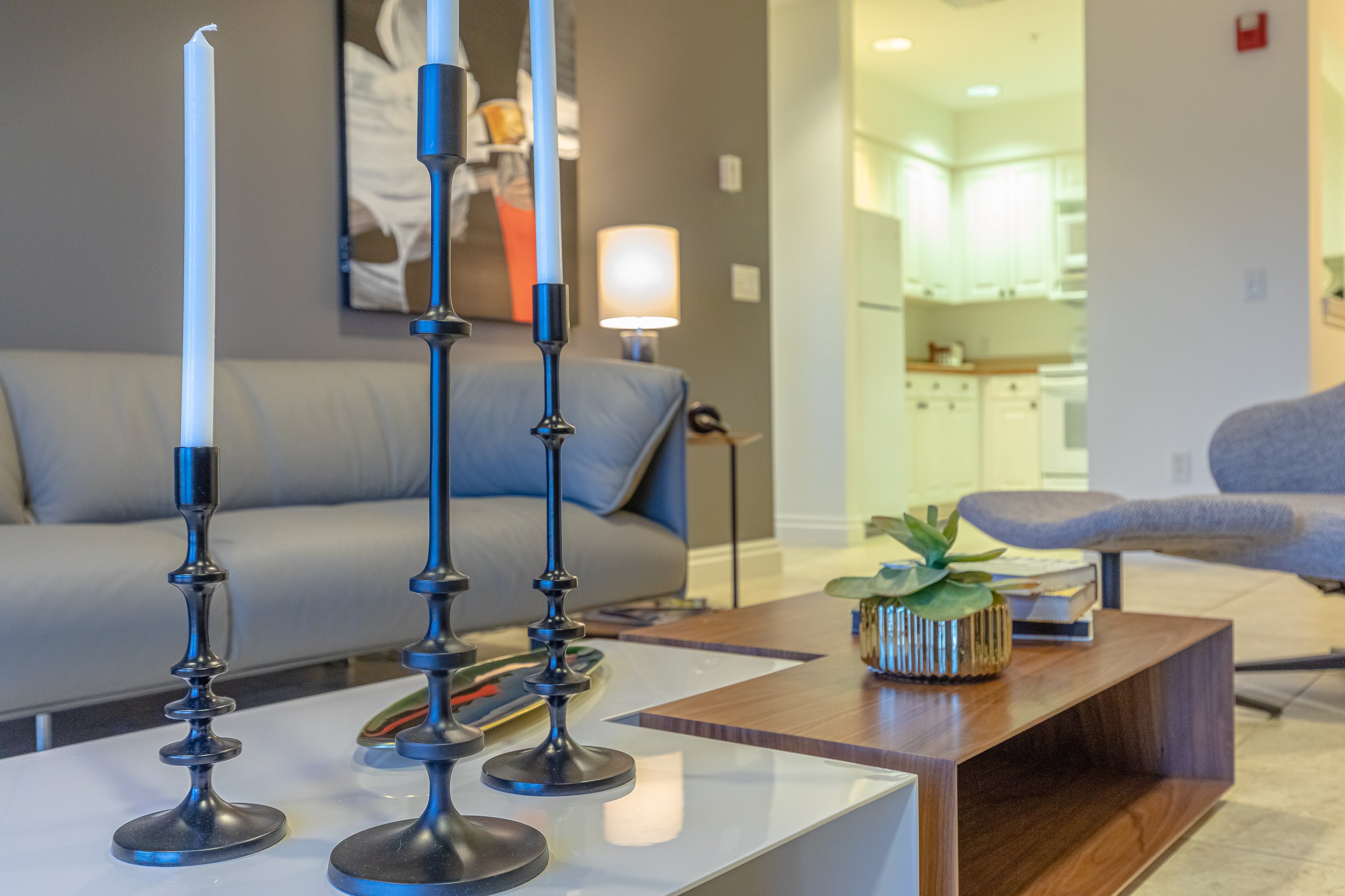 Living room with a white sofa, a wooden coffee table with a plant, modern candlesticks, and a gray chair. A lamp and artwork are on the walls, and a view into a kitchen with white cabinets is in the background.