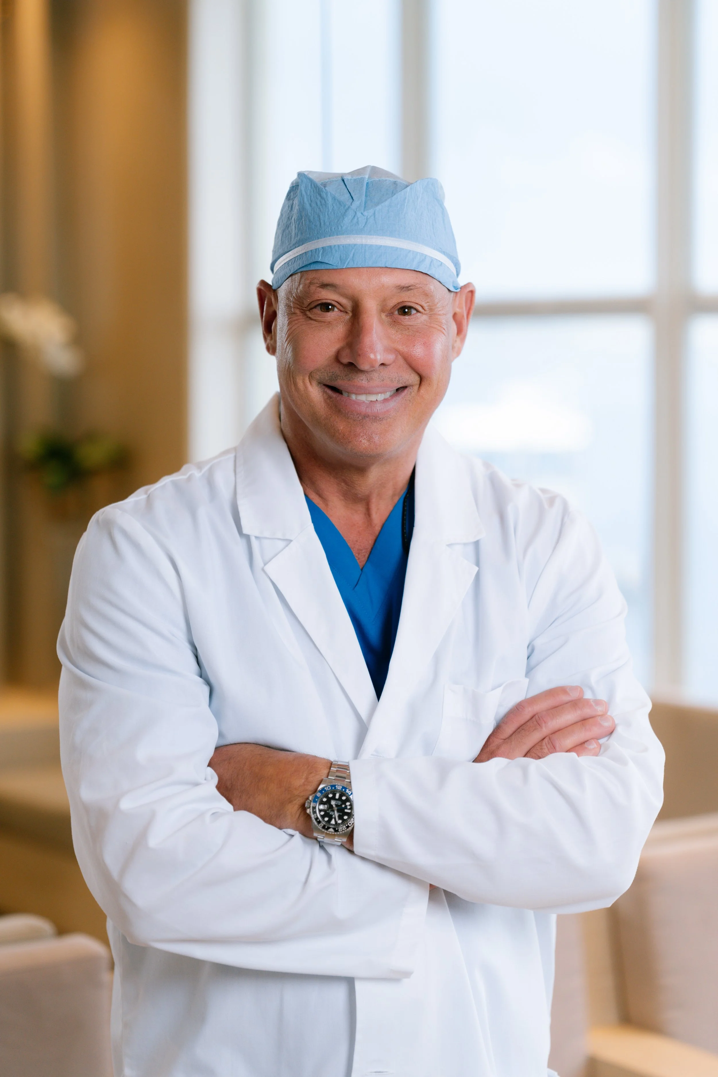 Smiling male doctor in white coat with crossed arms, wearing a blue surgical cap and watch, standing in a bright room.