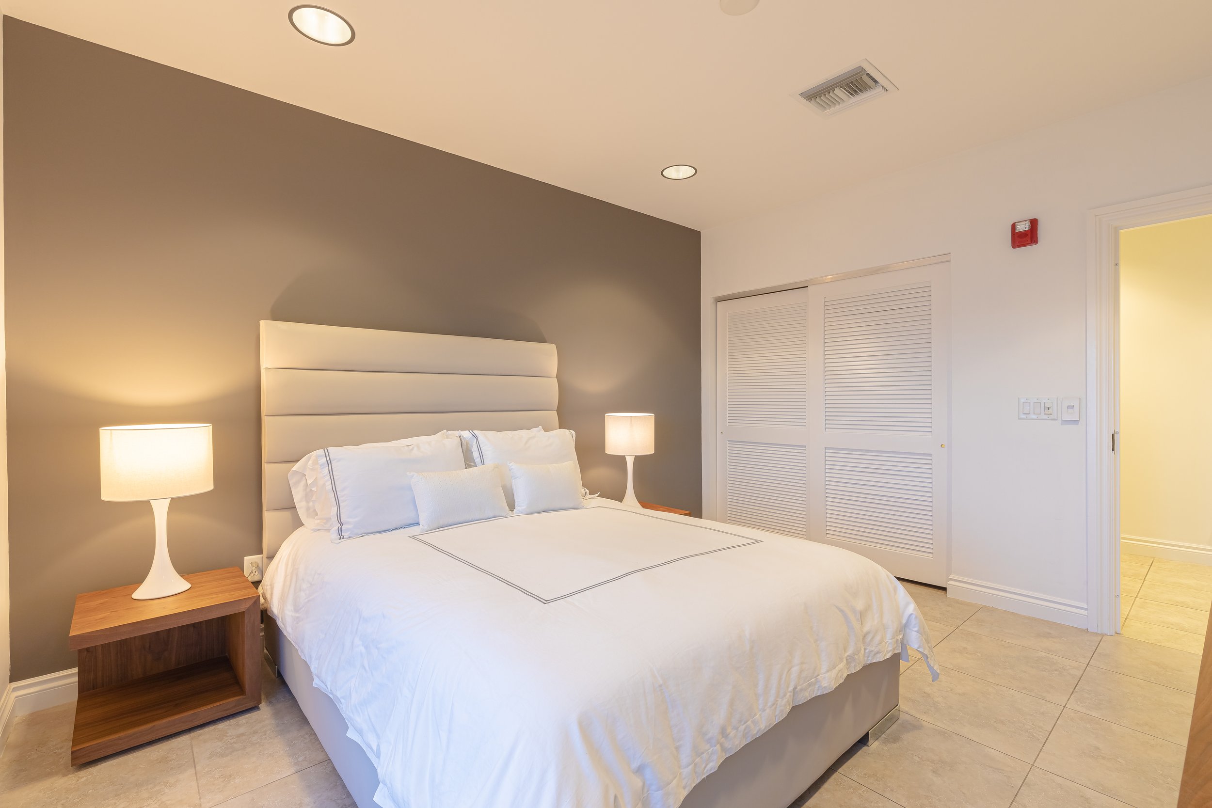 A bedroom with a bed, white bedding, pillows, two bedside lamps, a dark gray accent wall, a closet with louvered doors, and a doorway leading to a hallway.