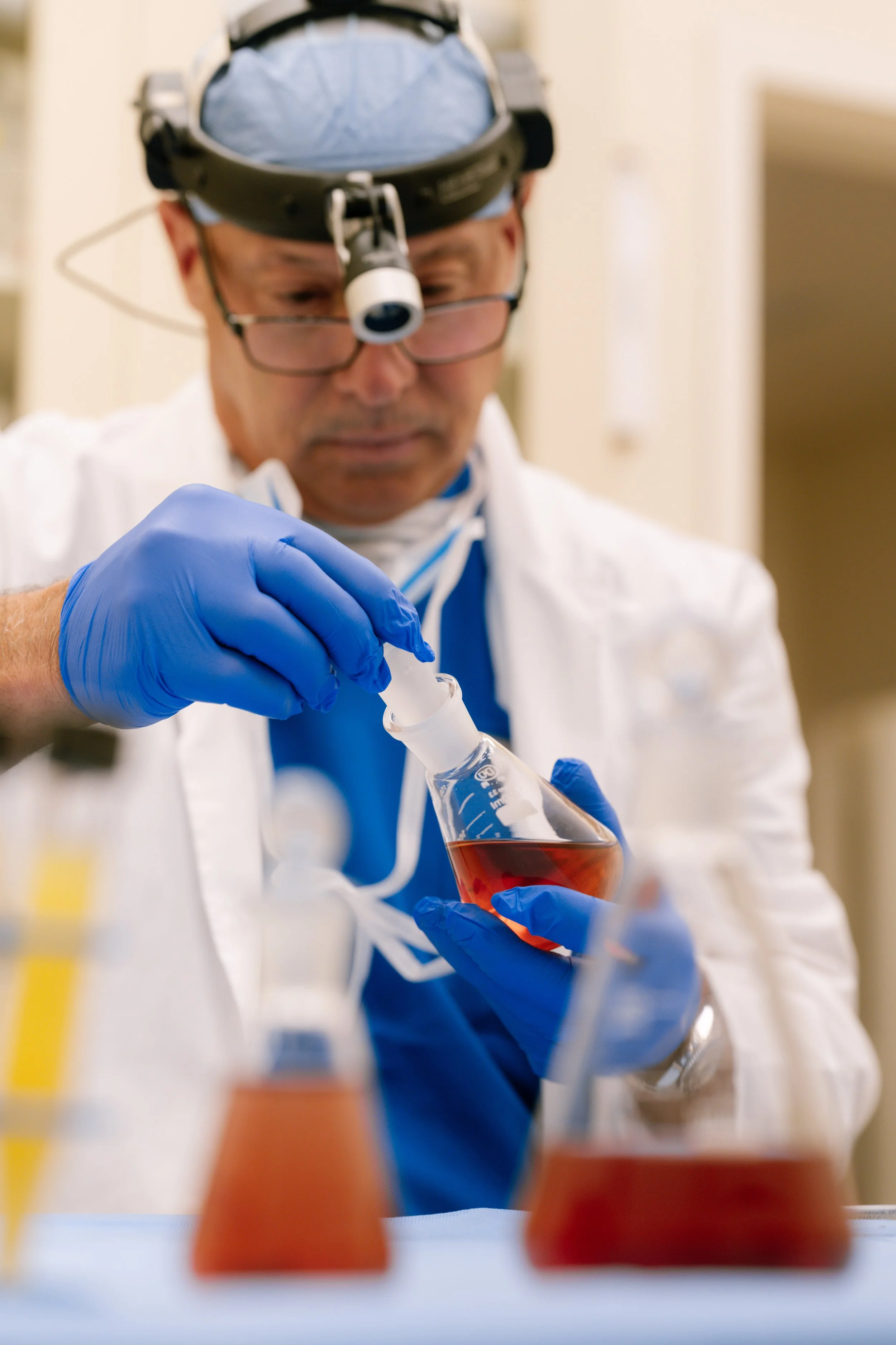 A scientist wearing a headlamp, glasses, blue gloves, and a white lab coat conducts an experiment with red liquid in laboratory glassware.