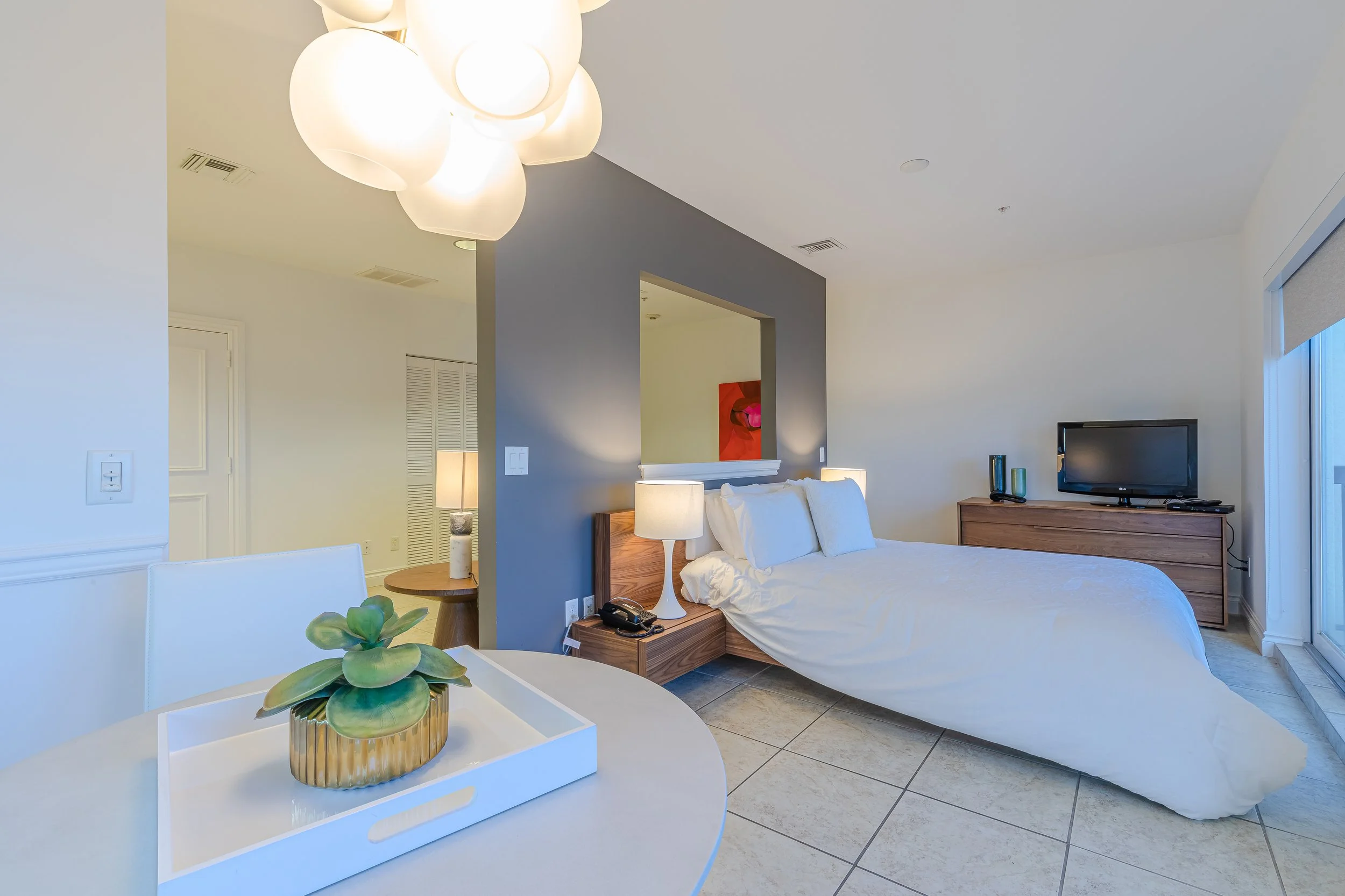 Interior of a modern suite with a bed, nightstand, dresser with television, dining table with a plant, and chandelier.