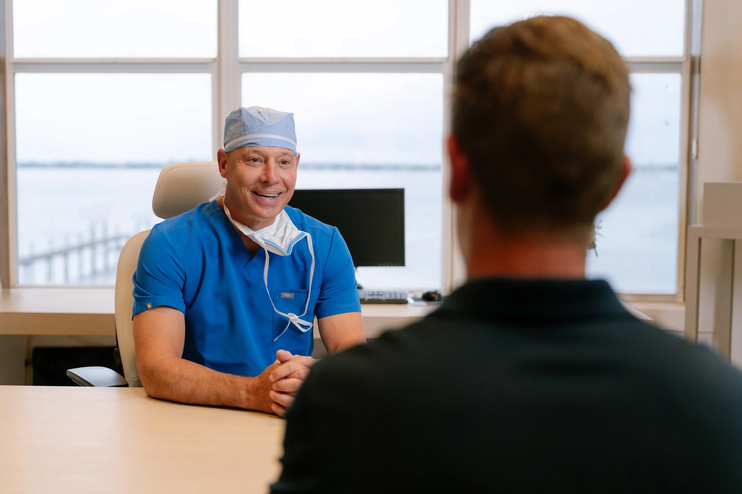 A doctor in blue scrubs and a surgical cap smiling and talking to a patient in an office with large windows showing water outside.