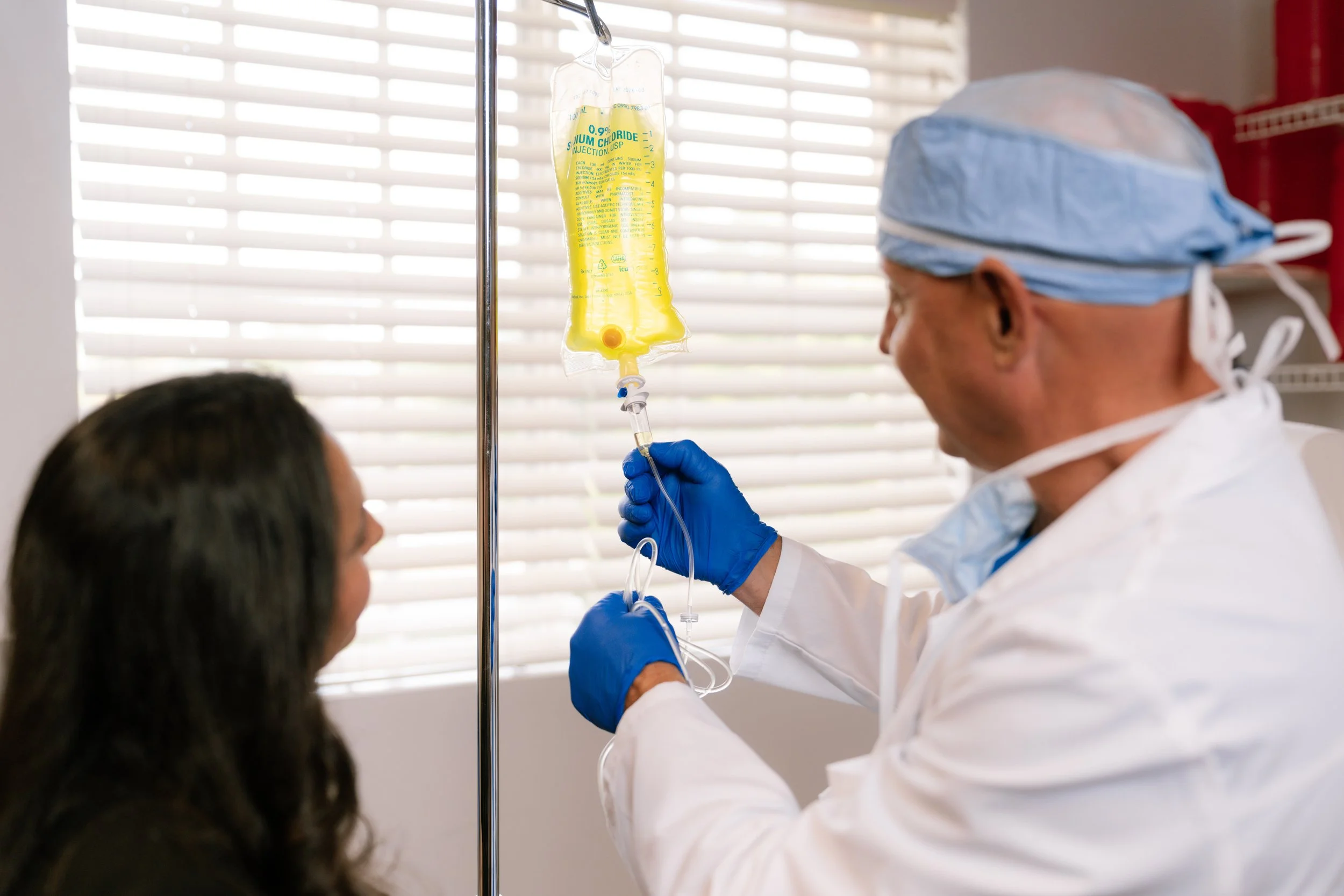A healthcare professional in blue gloves and scrubs administers an IV infusion to a patient in a medical setting.