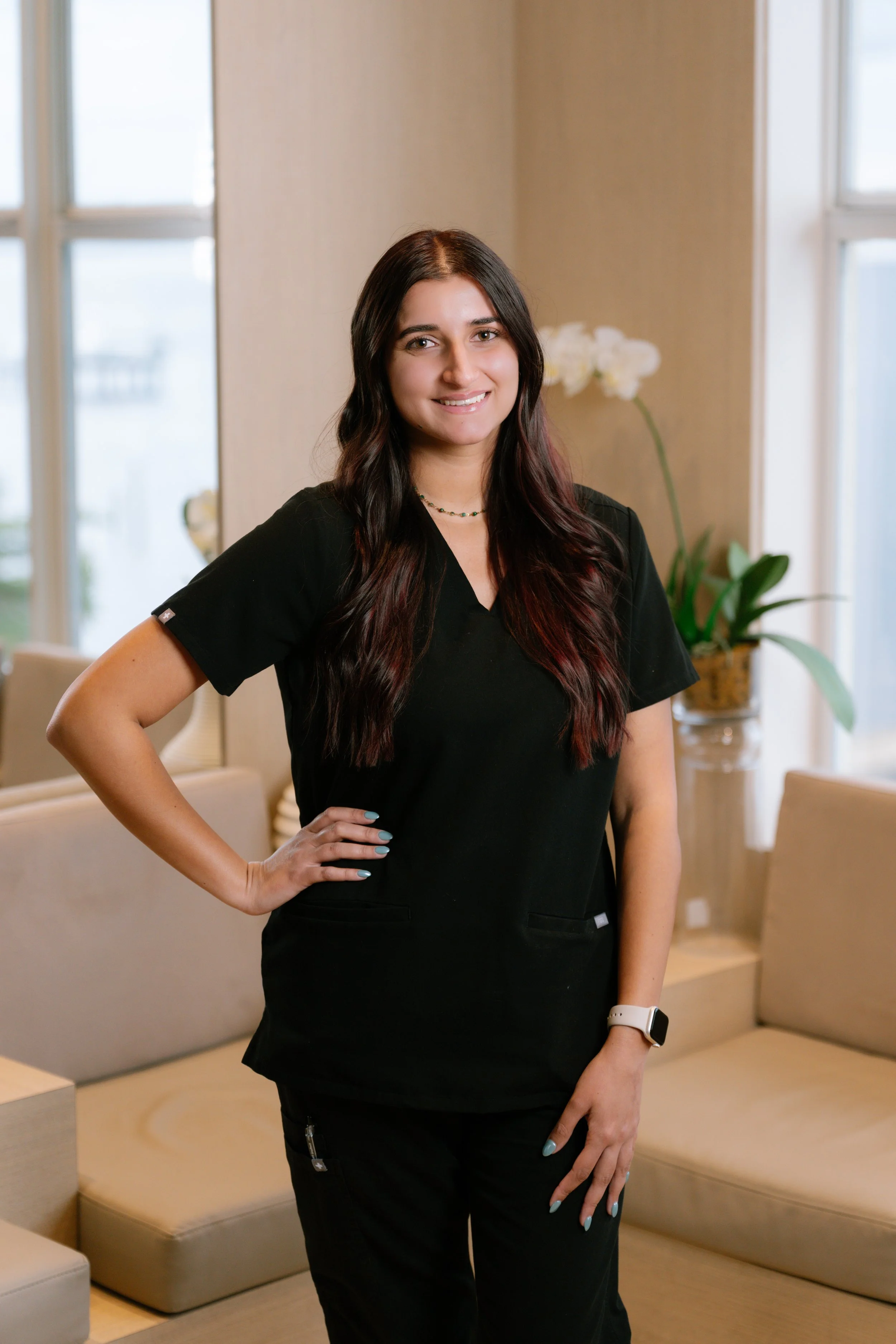 A woman with long dark hair, wearing a black short-sleeved shirt, standing with arms crossed inside a room with a window and a decorative vase in the background.