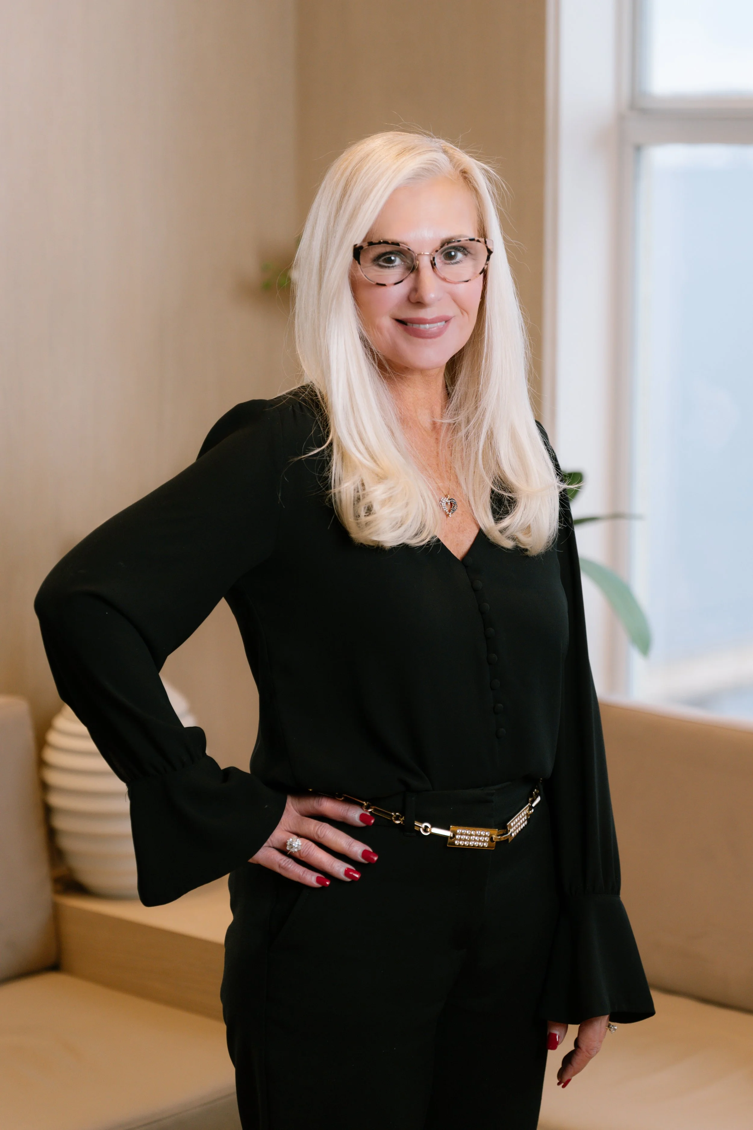 A professional woman with long blonde hair, glasses, wearing a black blouse and pants, standing indoors near a beige couch and large window.