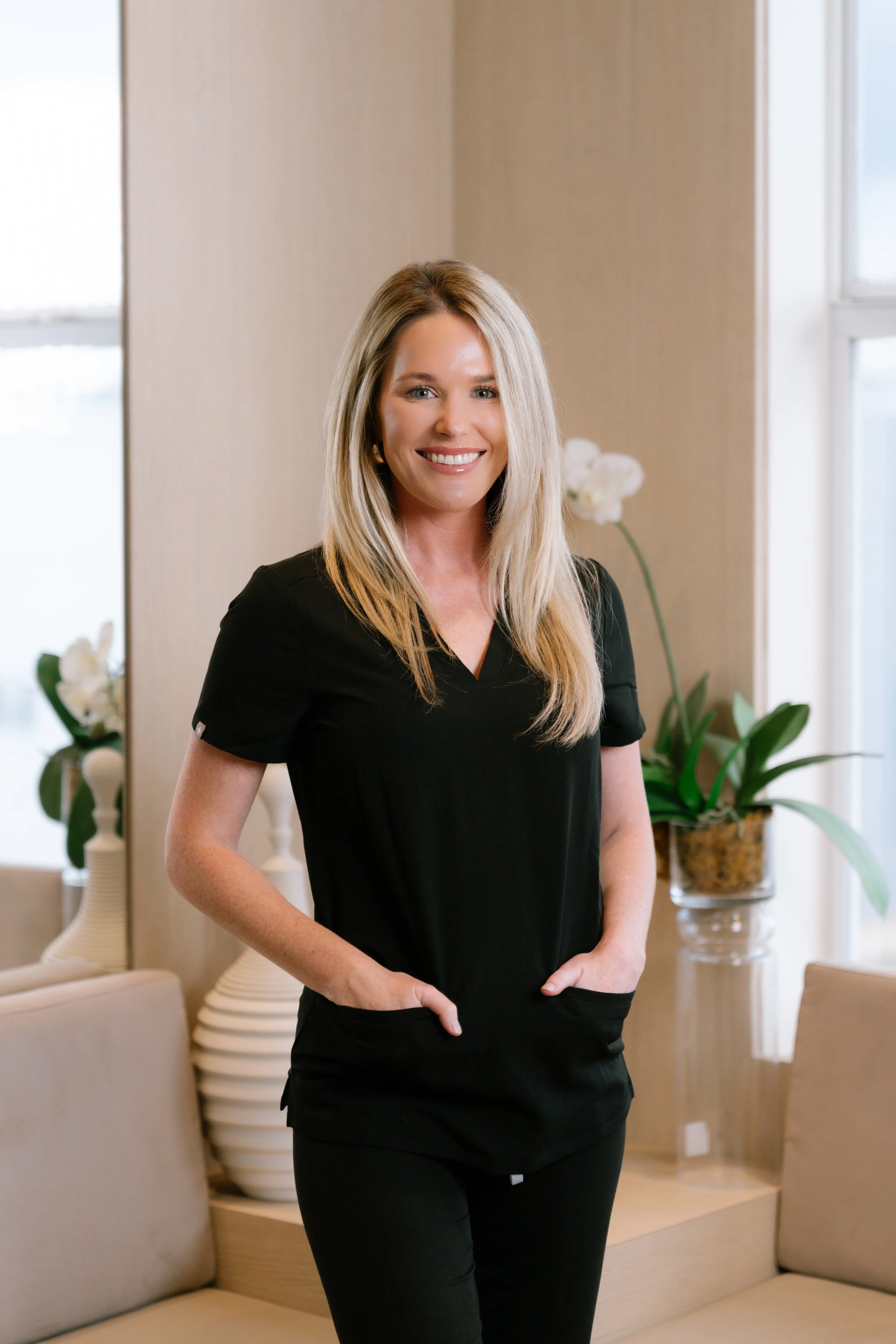A smiling blonde woman wearing a black V-neck top and black pants standing in a well-lit room with a mirror, beige walls, and decorative plants.