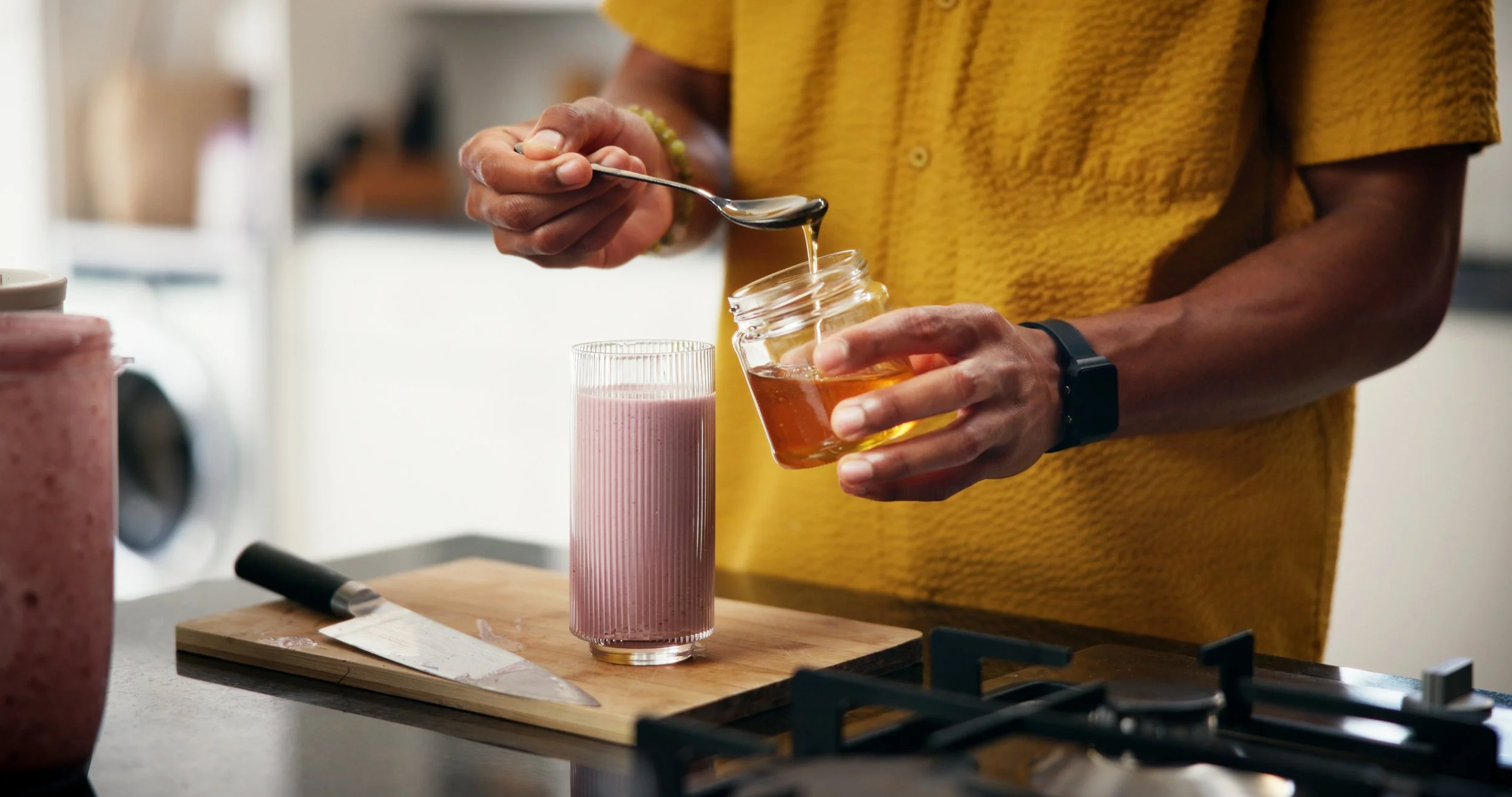 A person in a yellow shirt holds a jar of honey in one hand and a spoon in the other, drizzling honey into a tall pink smoothie glass on a wooden cutting board, with a kitchen background.