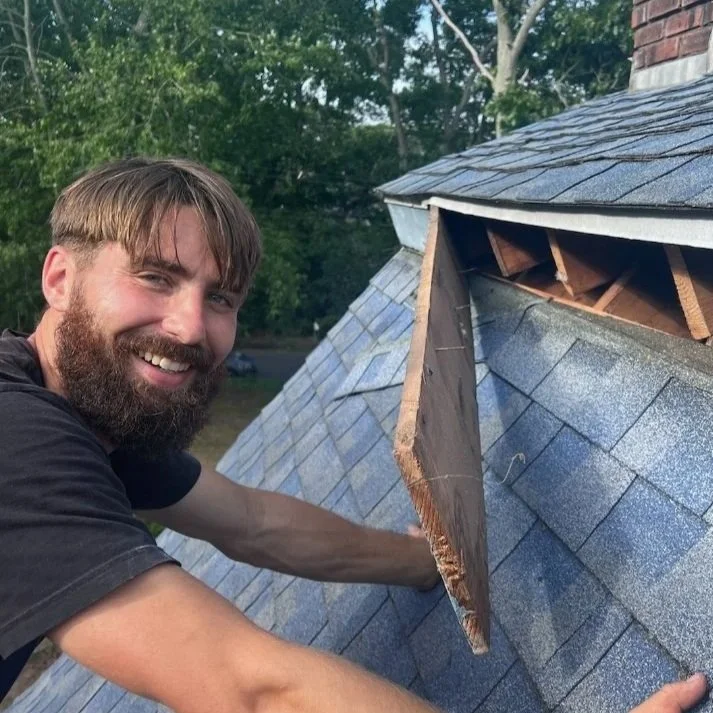 Man repairing or inspecting a roof with shingles, holding a broken piece of the roof's trim, standing outdoors on a sunny day.
