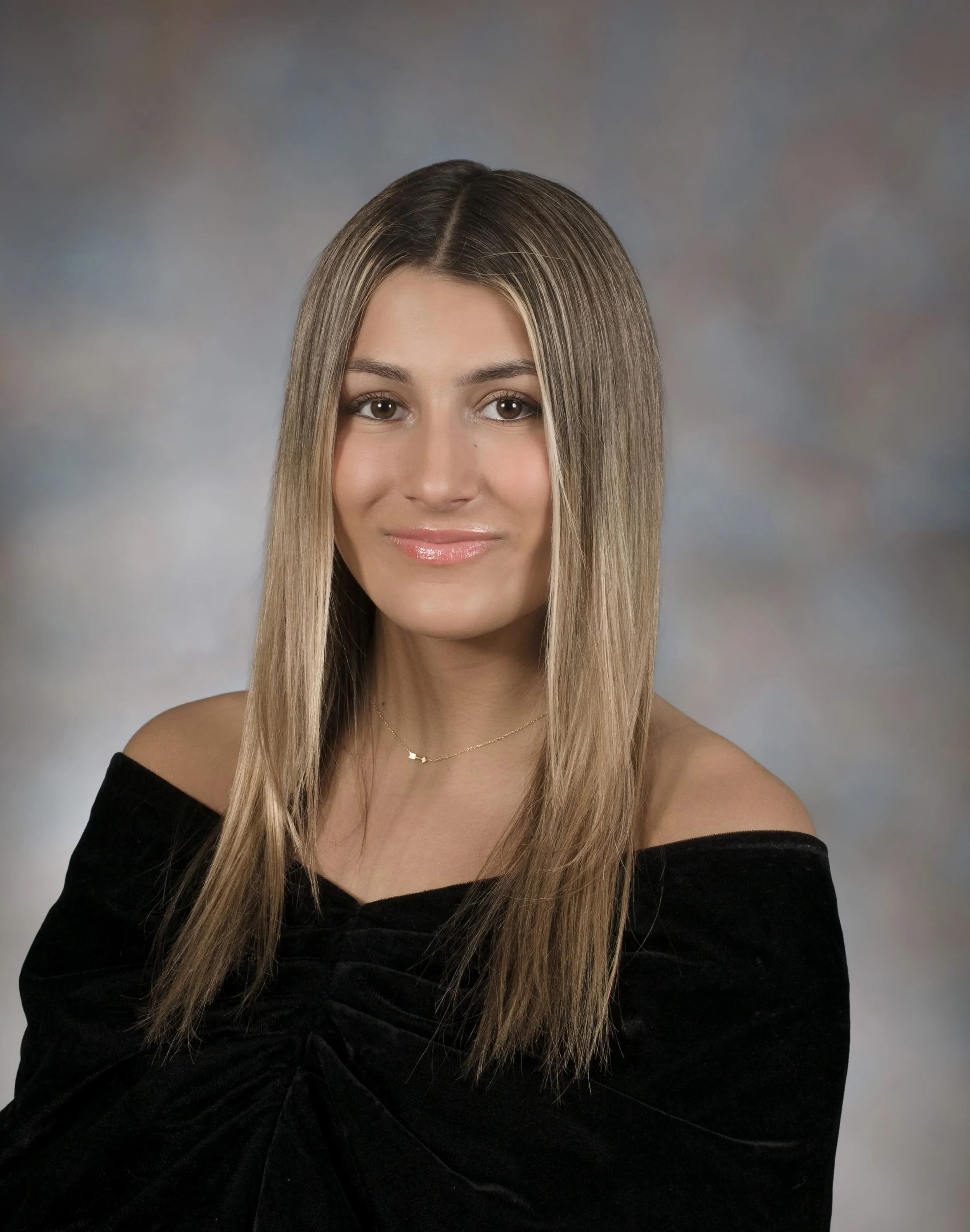 A young woman with long, straight blonde hair wearing an off-the-shoulder black top and a delicate gold necklace, smiling softly in front of a neutral, softly blurred background.
