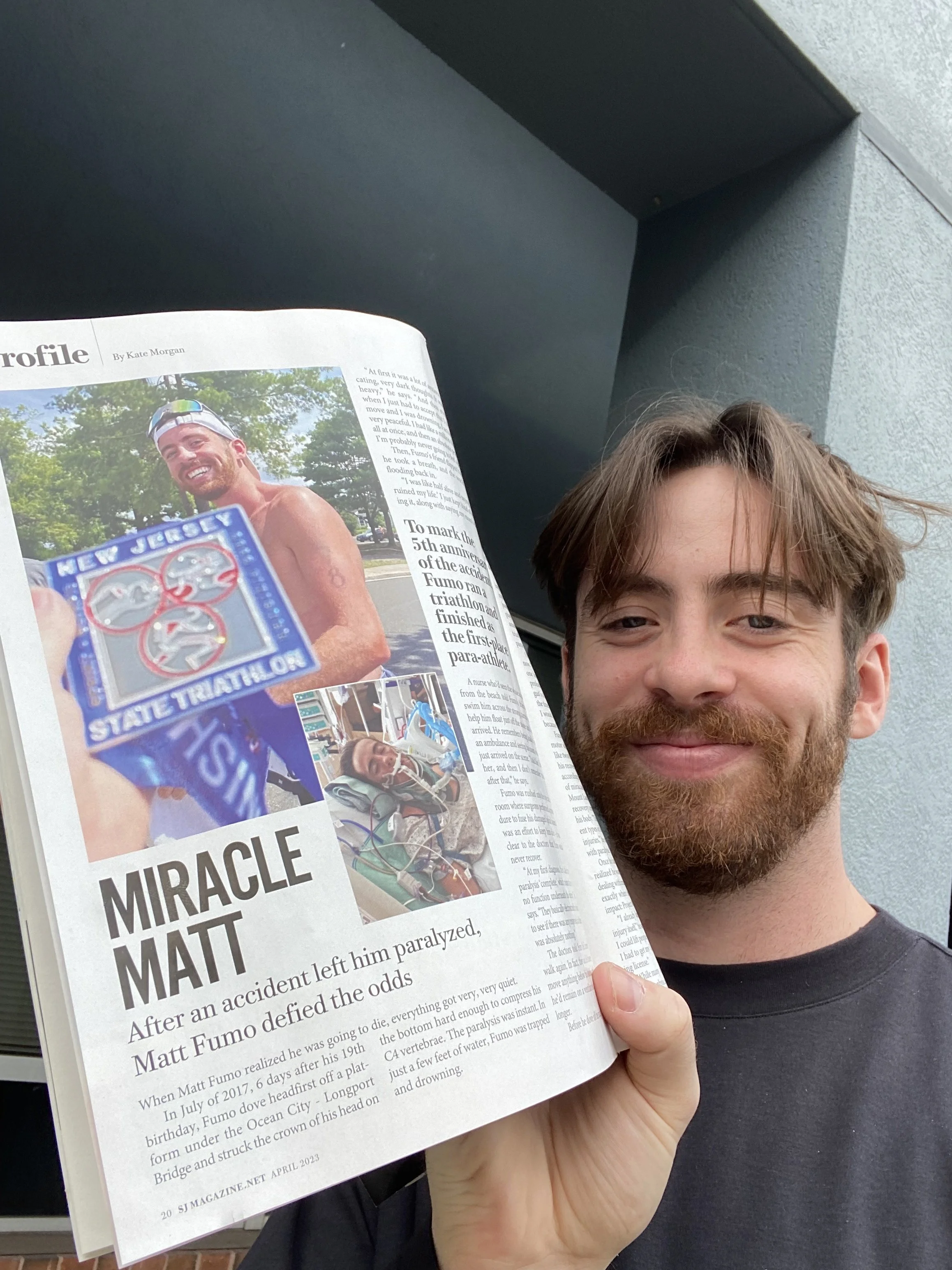 Smiling young man with brown hair and beard holding a magazine open, featuring a story about Matt Fumo, with a photo of Matt Fumo in a triathlon event.