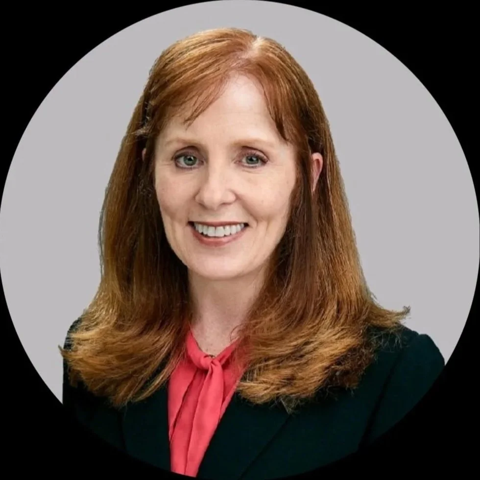 Headshot of a woman with red hair, smiling, wearing a dark blazer and a pink scarf against a gray background.