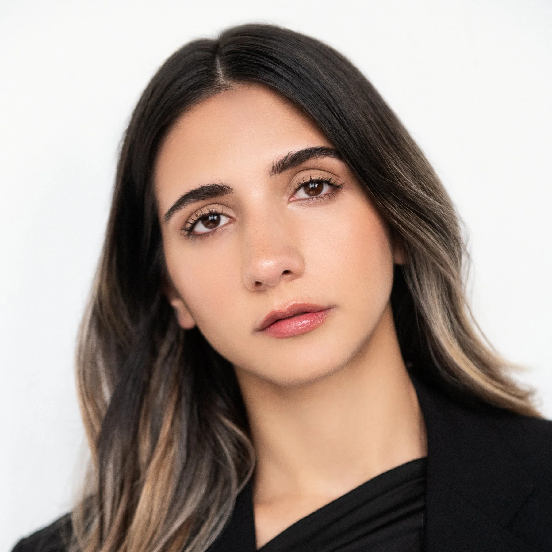 Portrait of a young woman with dark brown hair, light makeup, and a black blazer, looking directly at the camera against a white background.
