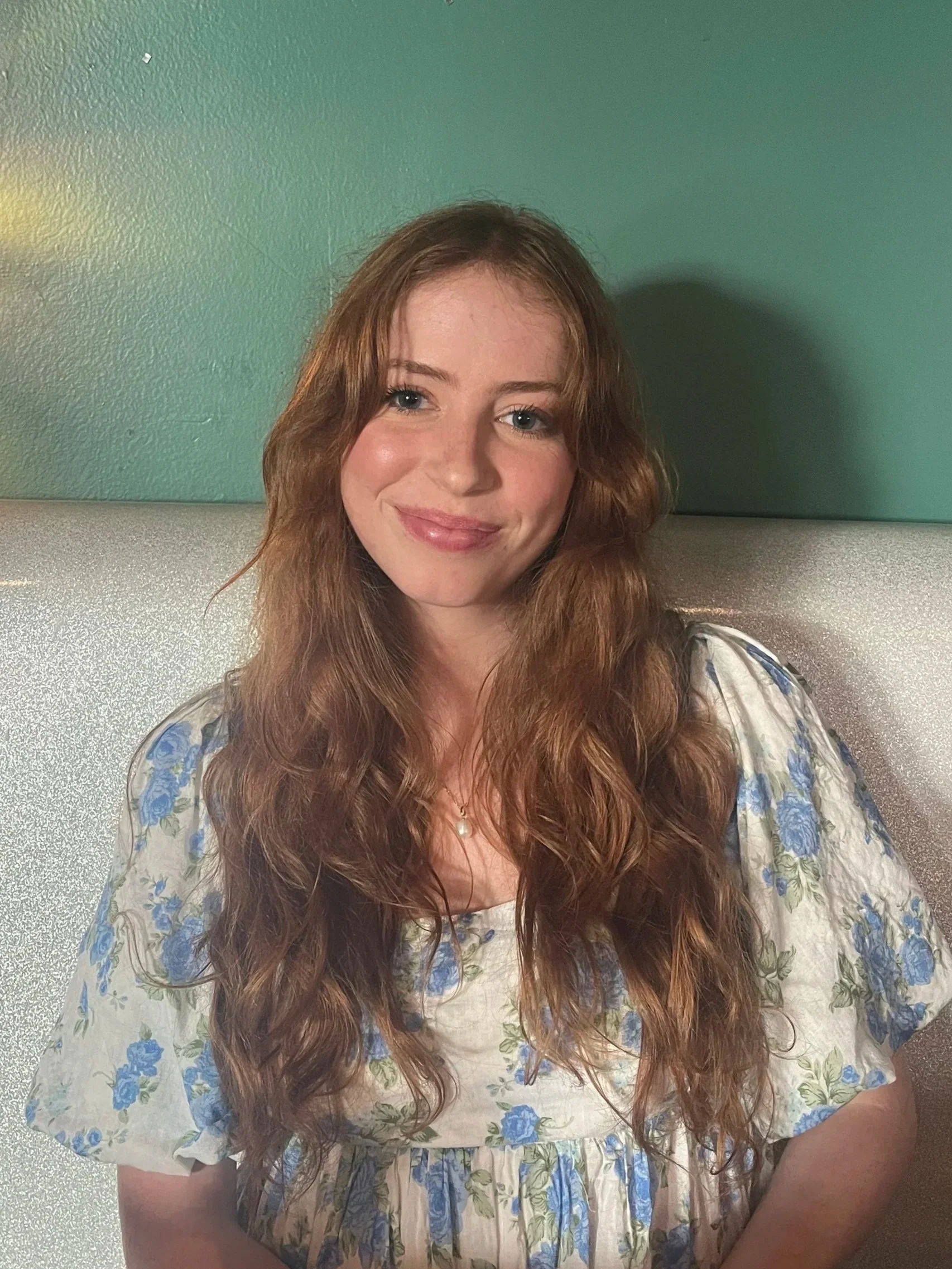 A young woman with long, wavy, auburn hair sitting against a green wall. She is smiling and wearing a cream-colored dress with blue floral patterns and a necklace.