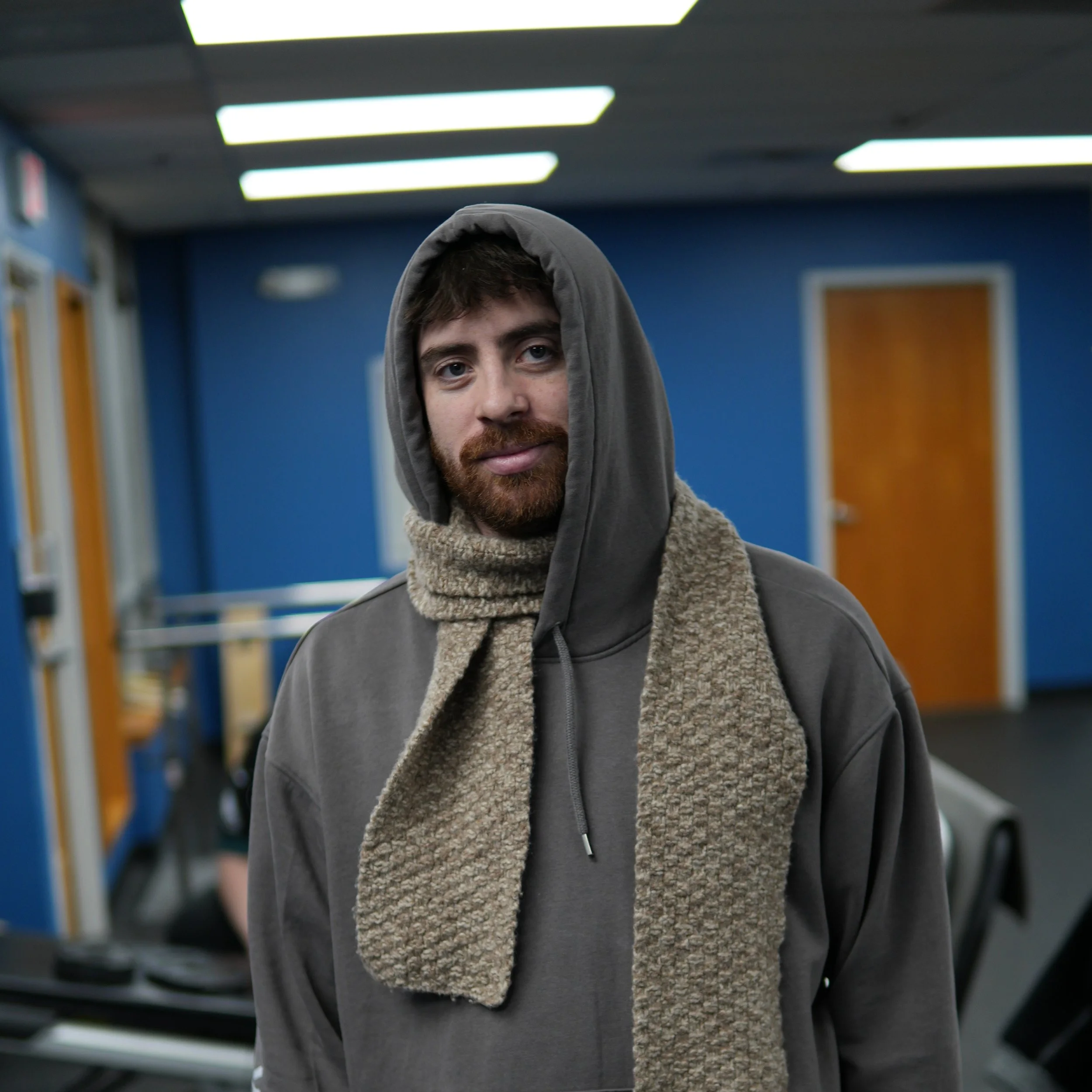 A man with a beard wearing a gray hoodie and a beige scarf, standing indoors with blue walls and fluorescent lighting.