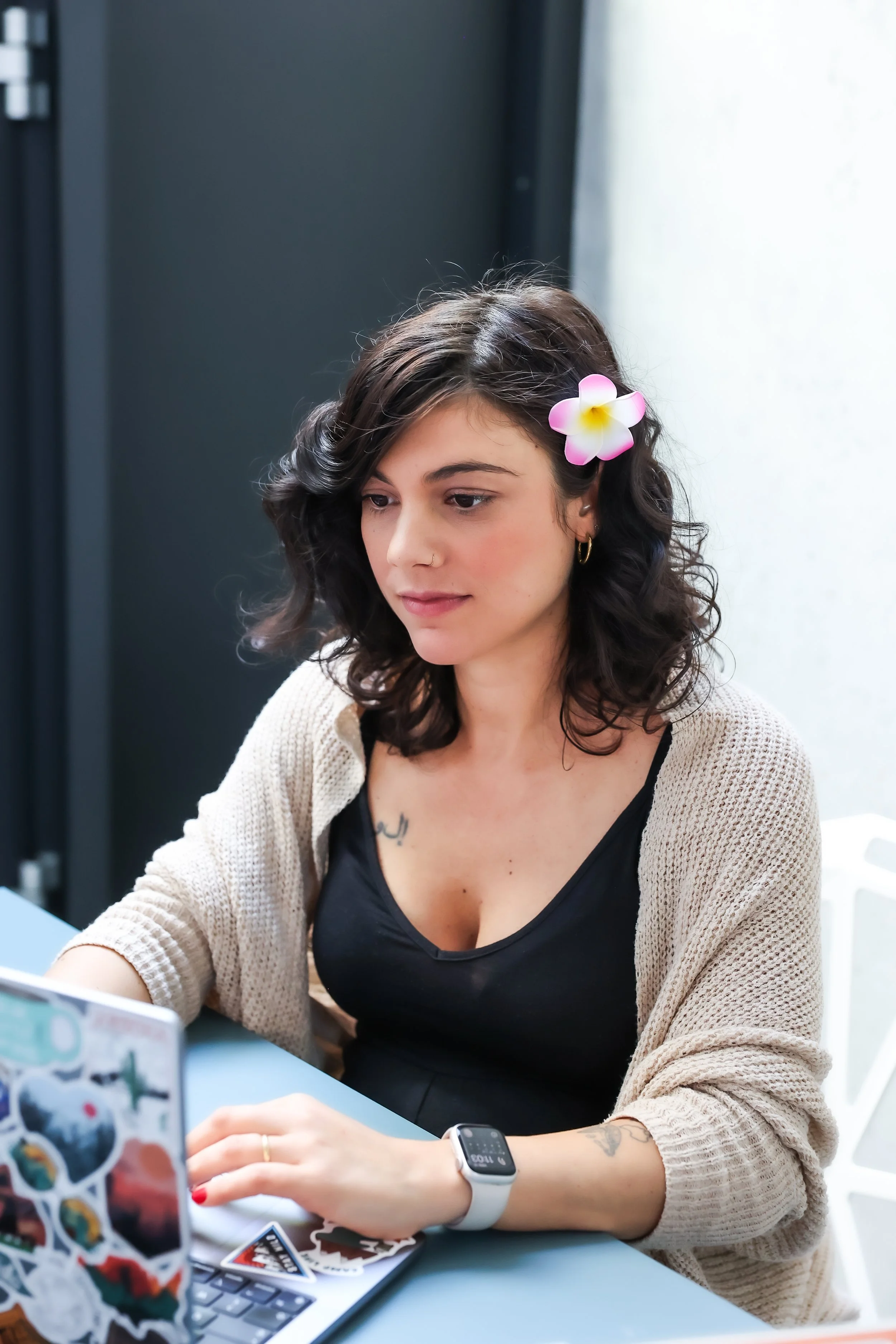 Jeune femme aux cheveux bruns, portant une fleur dans les cheveux, assise à une table, utilisant un ordinateur portable et portant une montre connectée qui tarifie des soins infirmiers aux mutuelles belges.