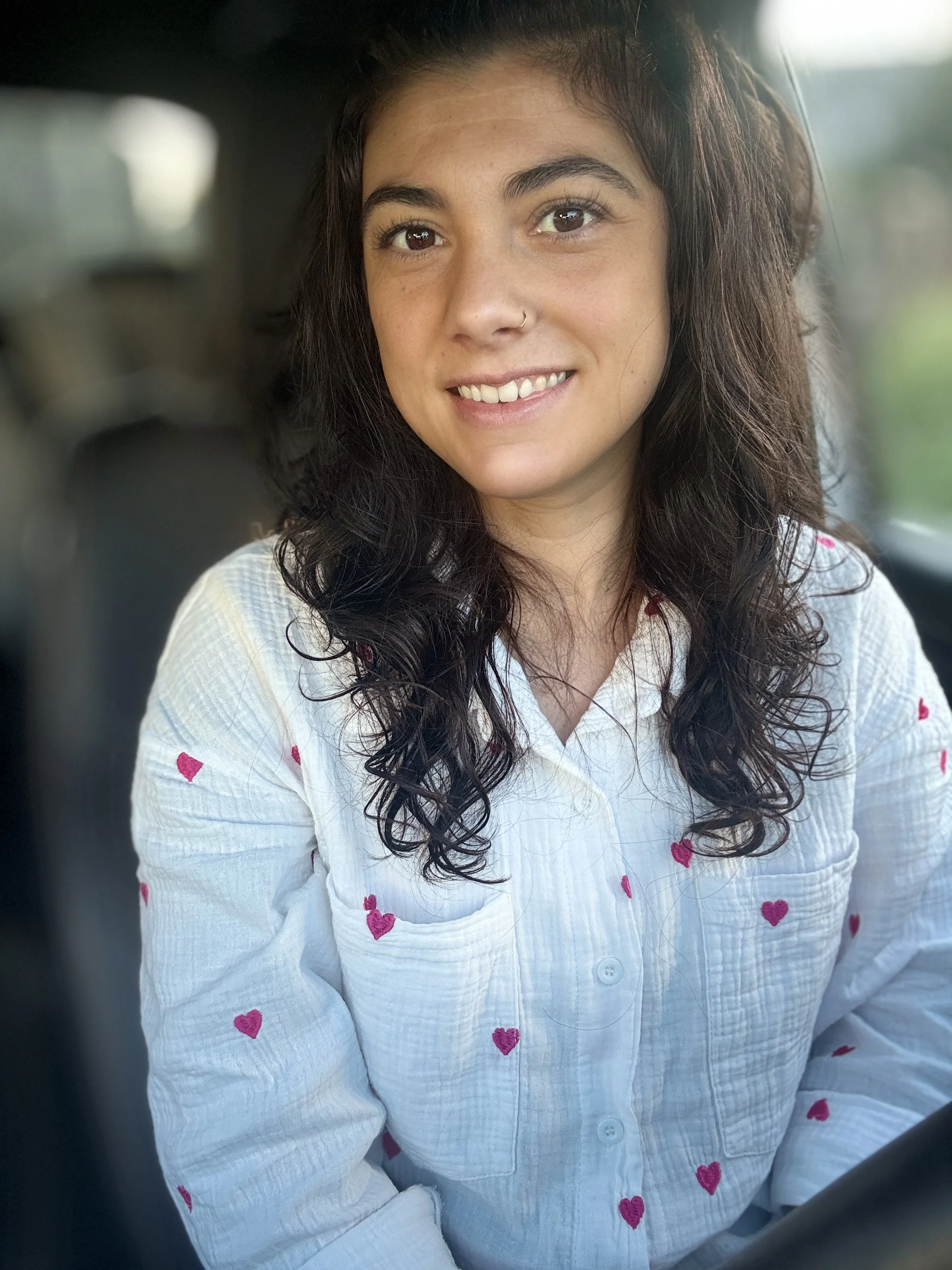 Une jeune femme souriante avec des cheveux bouclés, portant une chemise blanche avec des cœurs roses, assise probablement dans une voiture pendant sa tournée en soins à domicile.