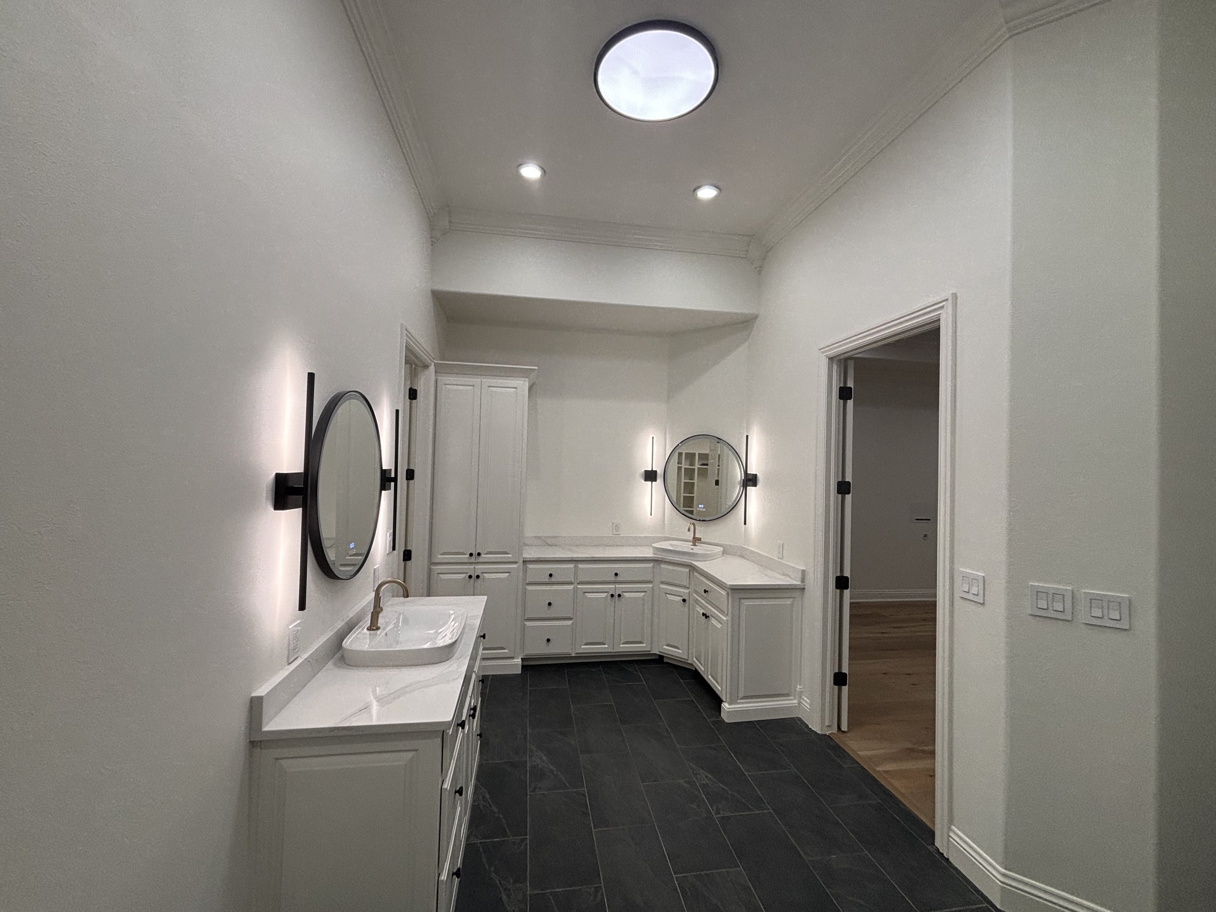 Empty bathroom with white cabinets, two round mirrors, black wall lights, and dark tile flooring.