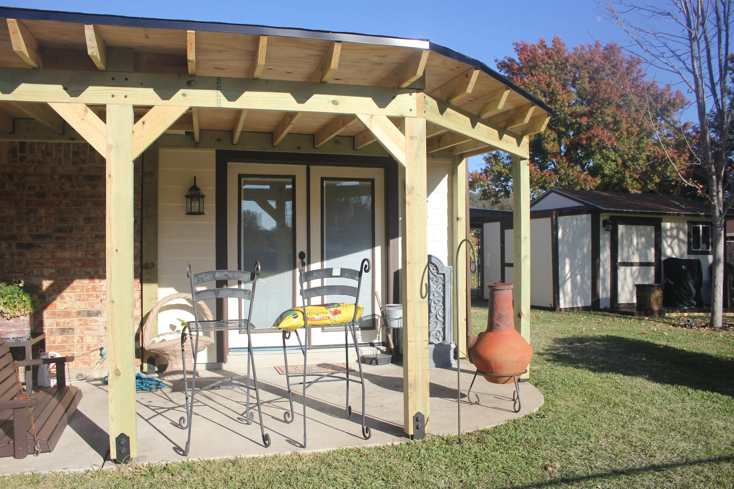 Back porch of a house under construction with a wooden frame, patio furniture, a chiminea, and a shed in the background, surrounded by grass and trees.