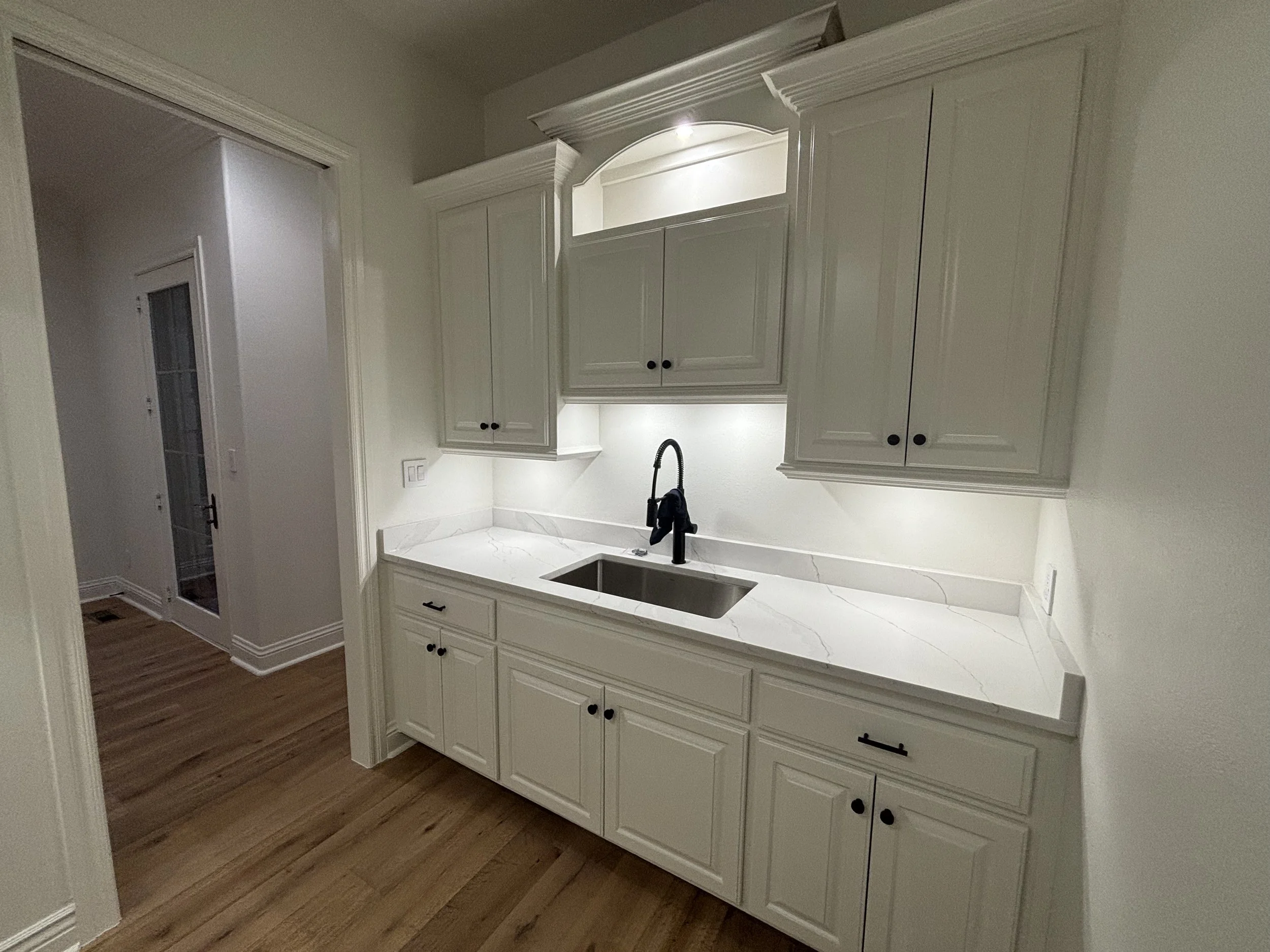 White kitchen cabinet and countertop with a sink and black faucet, under cabinet lighting, next to a doorway with hardwood floors.