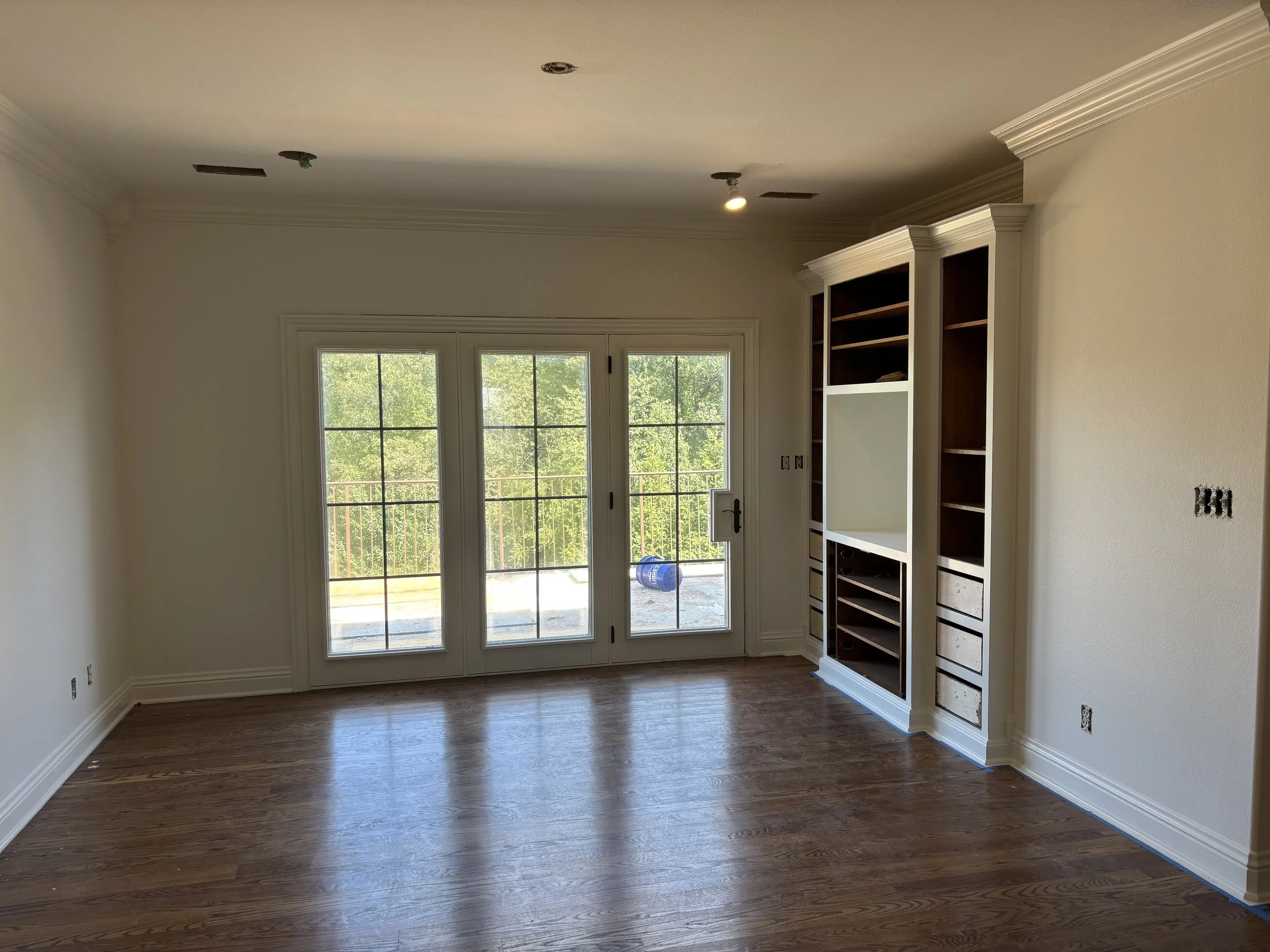 Empty living room with hardwood floor, white walls, large glass doors leading outside, built-in white and dark wood shelving unit, and ceiling light fixtures.