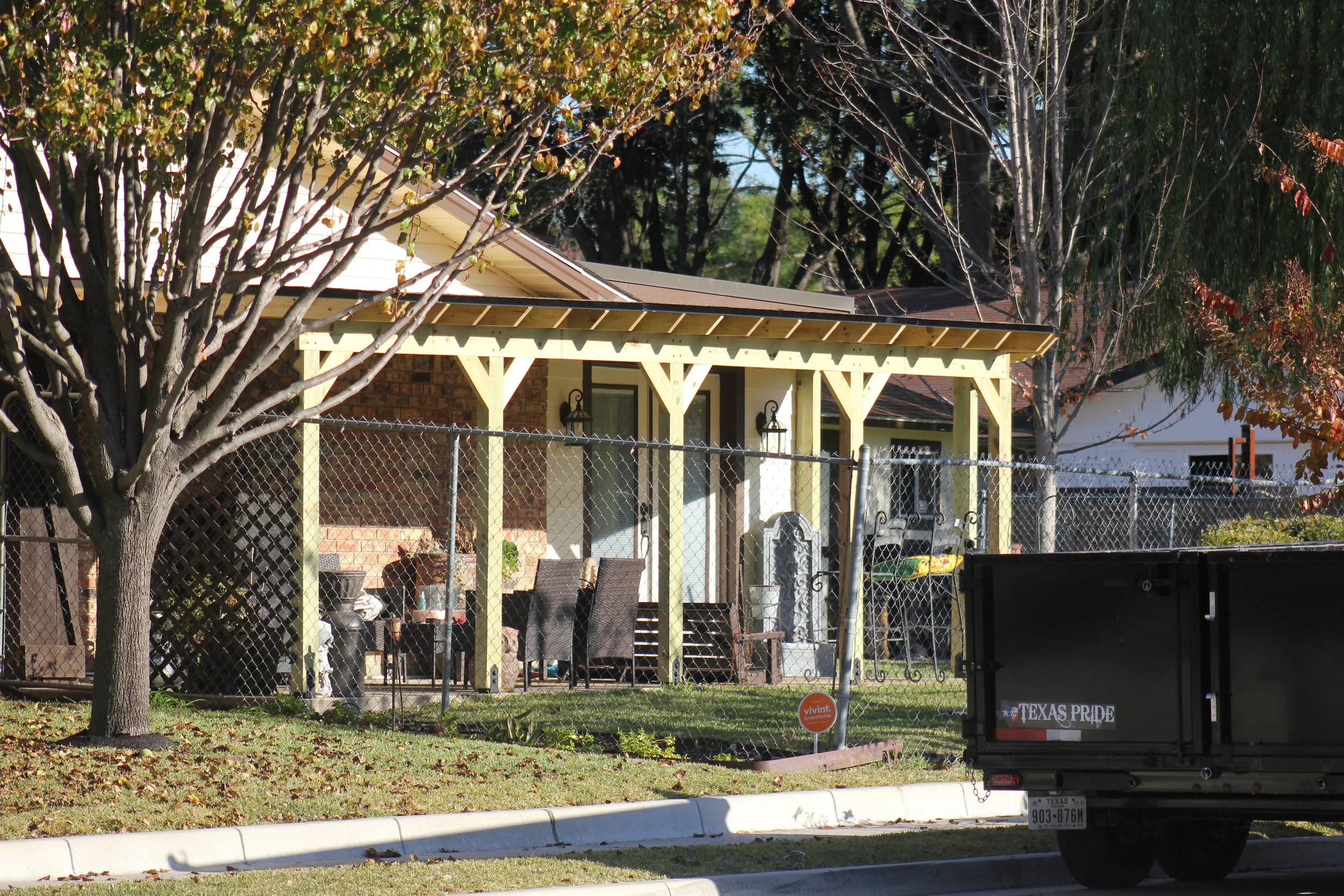A backyard with a covered patio, outdoor furniture, trees, a chain-link fence, and a truck labeled Texas Pride.