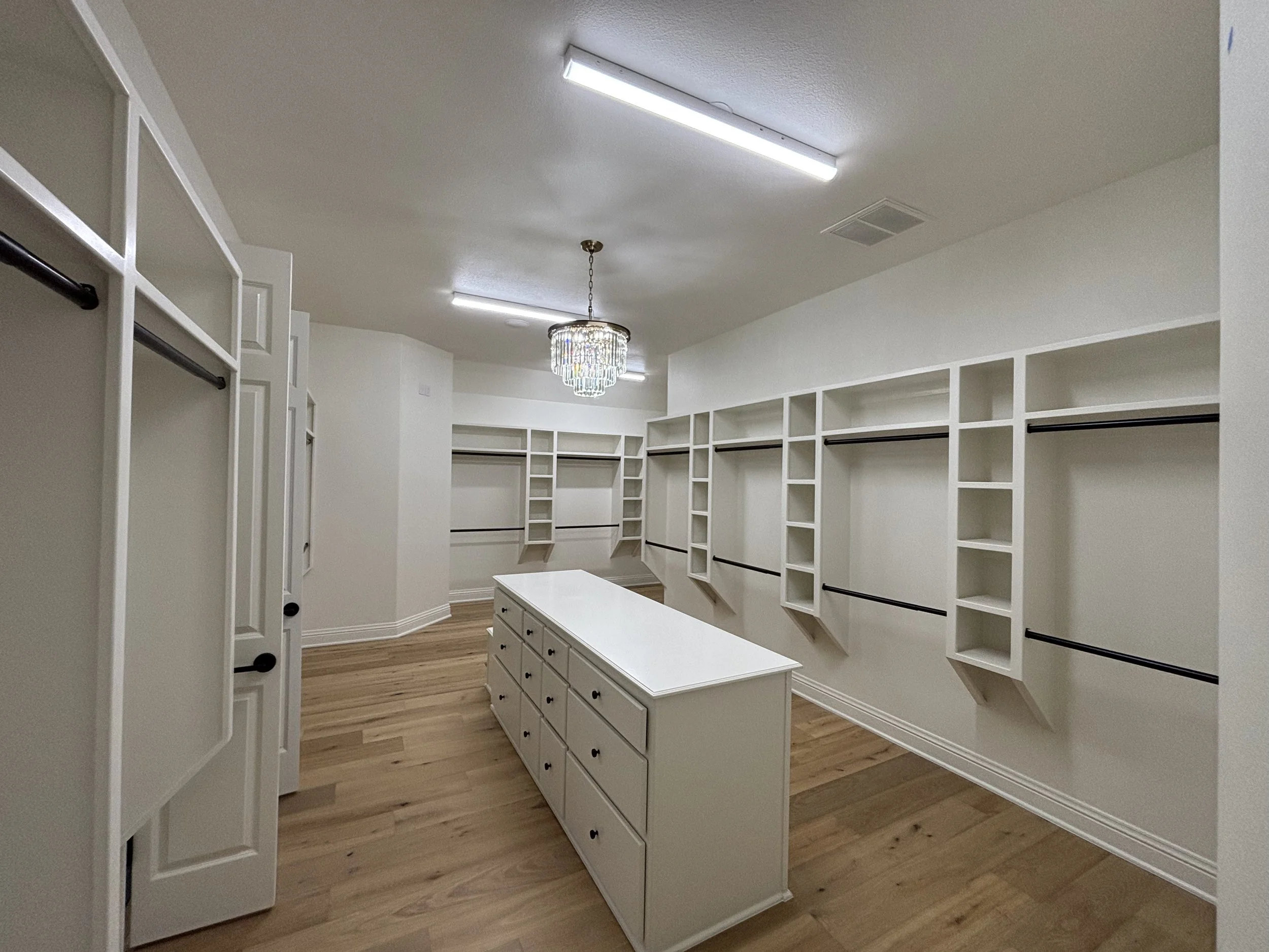 Empty walk-in closet with white shelves, black hanging rods, a central white drawer island, wood flooring, and ceiling lights, including a chandelier.