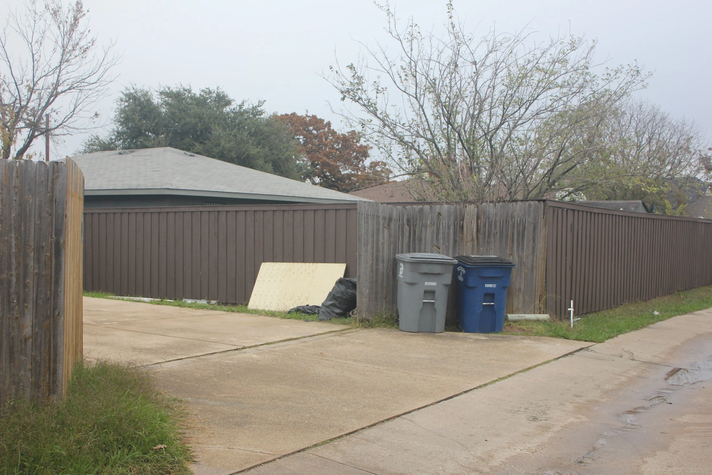 View of a backyard with a wooden fence, two trash bins, a discarded piece of furniture, and a wet driveway, on an overcast day.