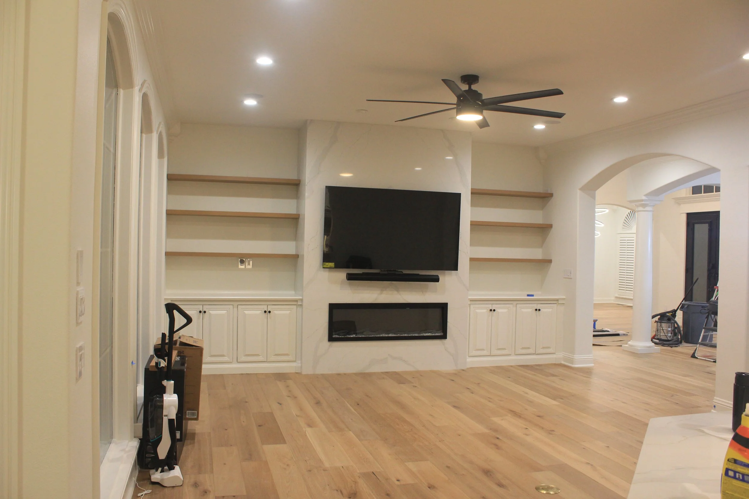 Living room with mounted TV, electric fireplace below, wooden shelves on either side, ceiling fan, hardwood floor, and partially visible furniture and tools.