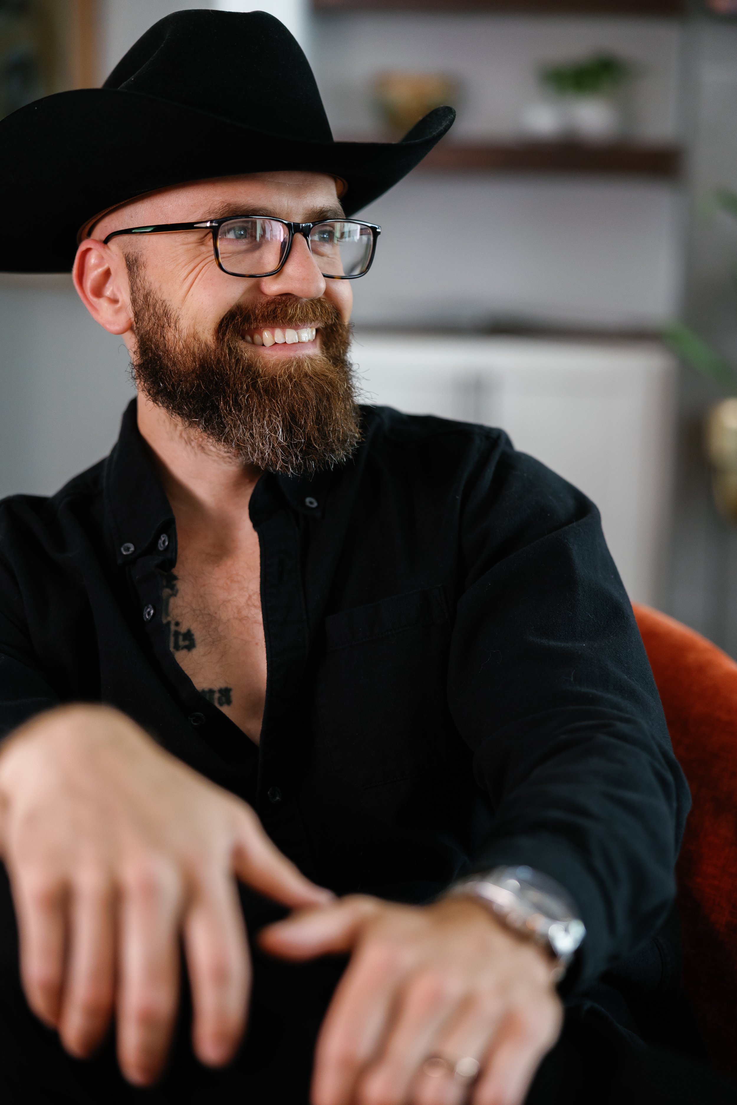 A smiling man with a beard, glasses, and a black cowboy hat, wearing a black shirt and a watch, sitting on a red chair in a cozy indoor setting.