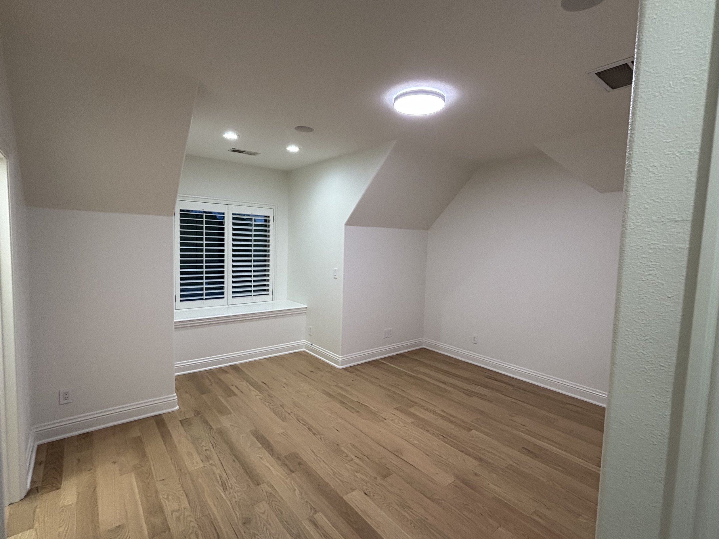 Empty room with white walls, hardwood floors, a window with white shutters, and ceiling lights.