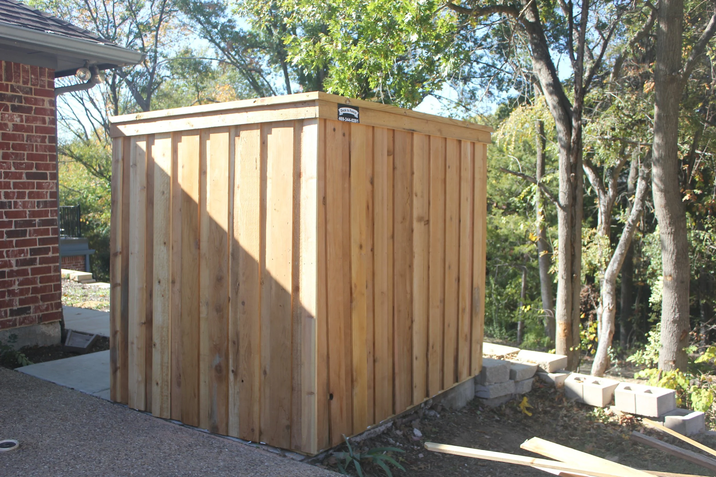 A newly built wooden shed attached to a brick house, surrounded by trees and some construction materials on the ground.