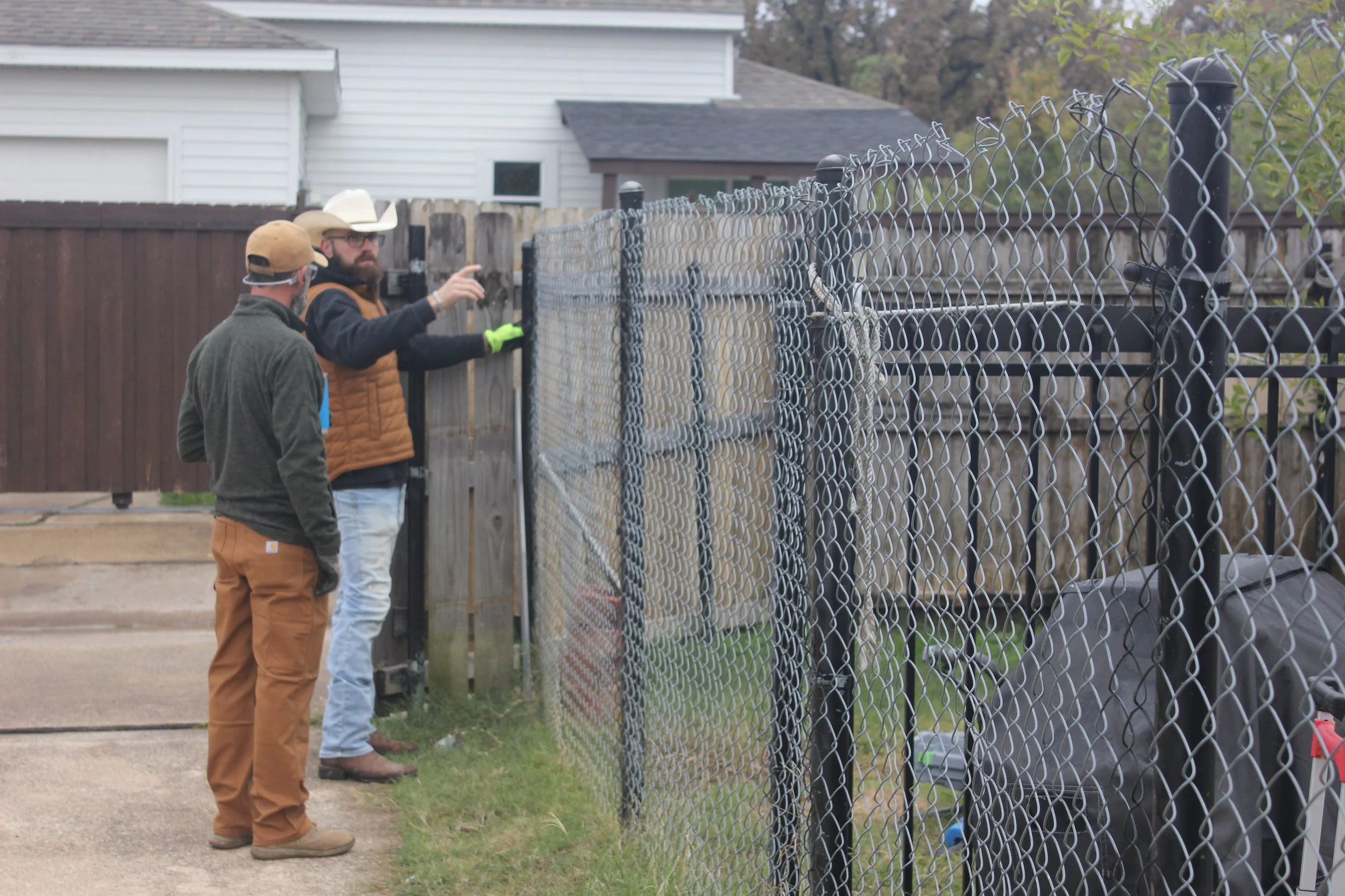 Two men are working on a chain-link fence. One man is pointing at something, while the other looks on. They are outdoors near residential houses.