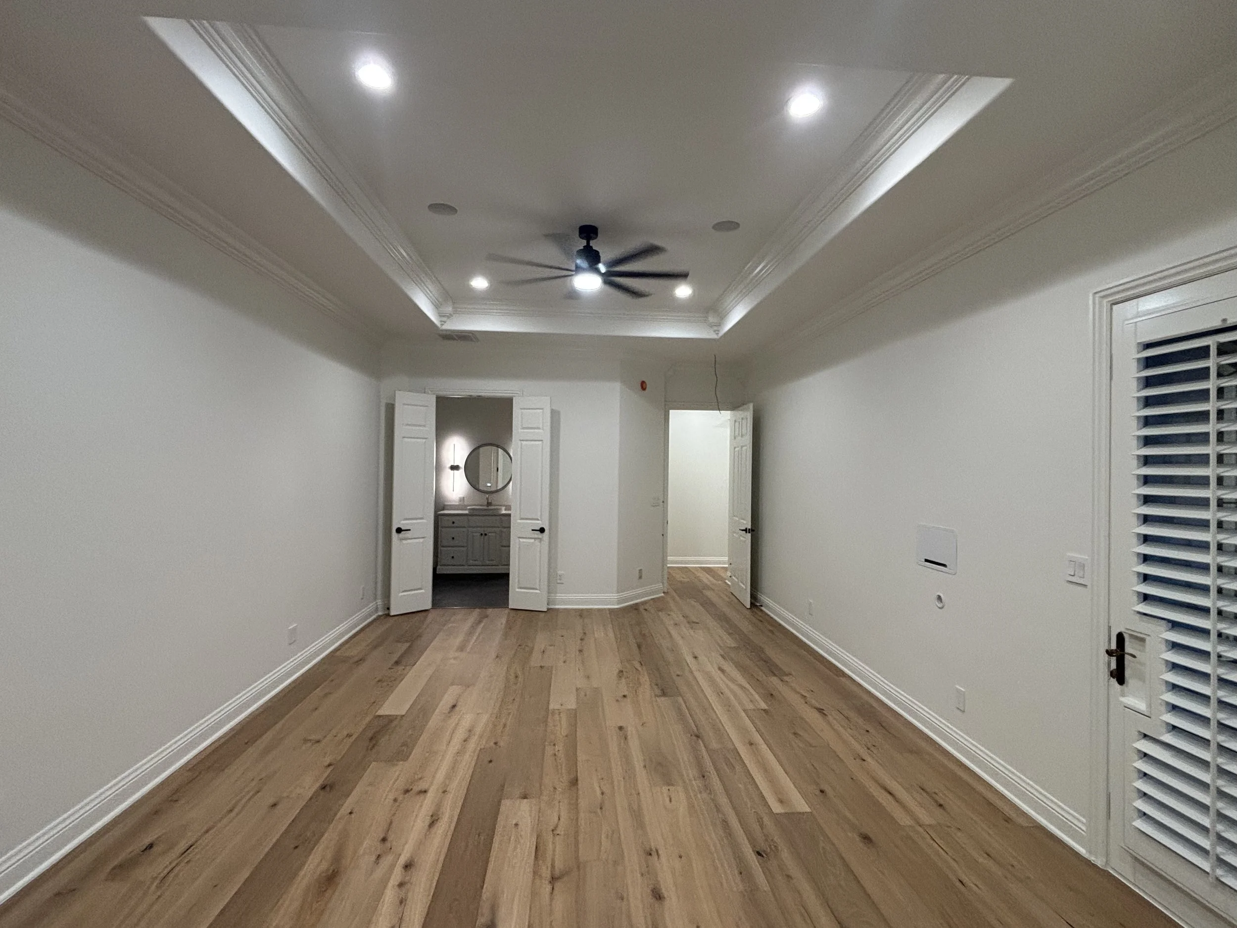 Empty living room with hardwood floors, white walls, white crown molding, a ceiling fan, recessed lighting, and a door leading to a bathroom with a mirror and vanity.