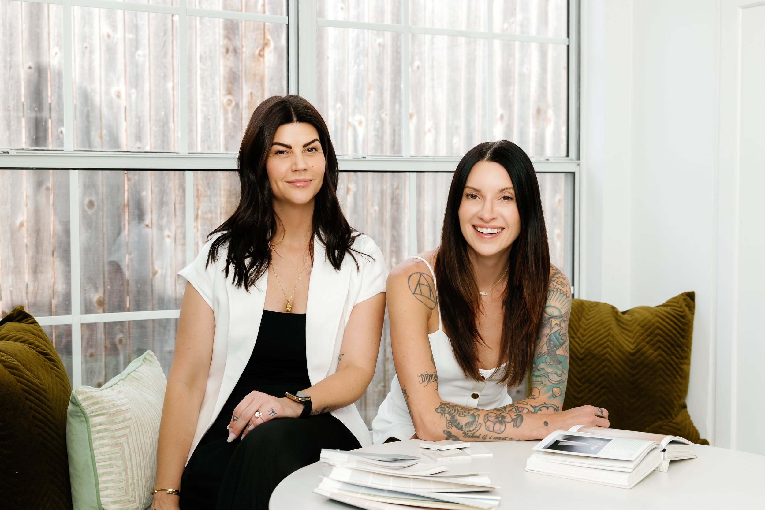 Two women sitting on a white couch in front of a large window, with magazines or papers on a white table in front of them, smiling at the camera.