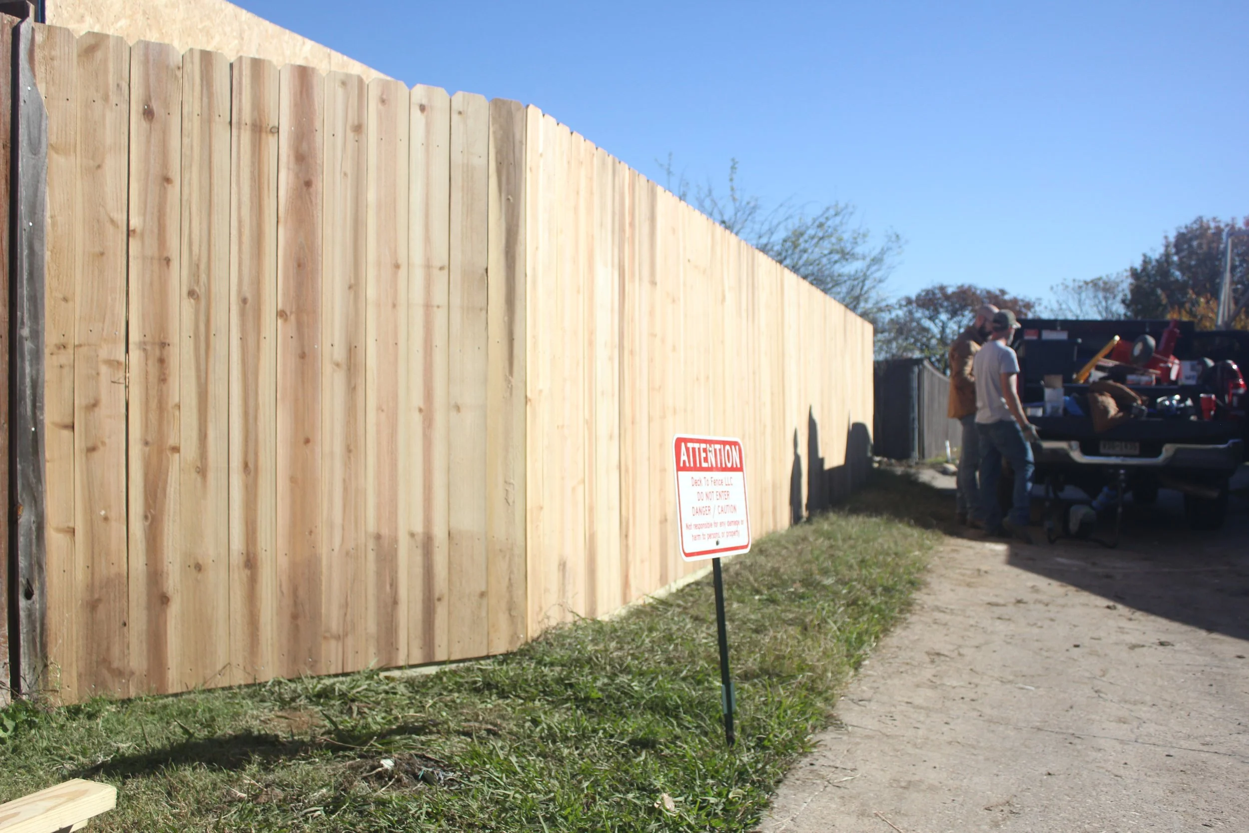 A wooden privacy fence alongside a dirt path with a red and white caution sign that reads "Attention" in front of the fence. In the background, three men are working near a pickup truck with tools and equipment in its bed, under a clear blue sky.