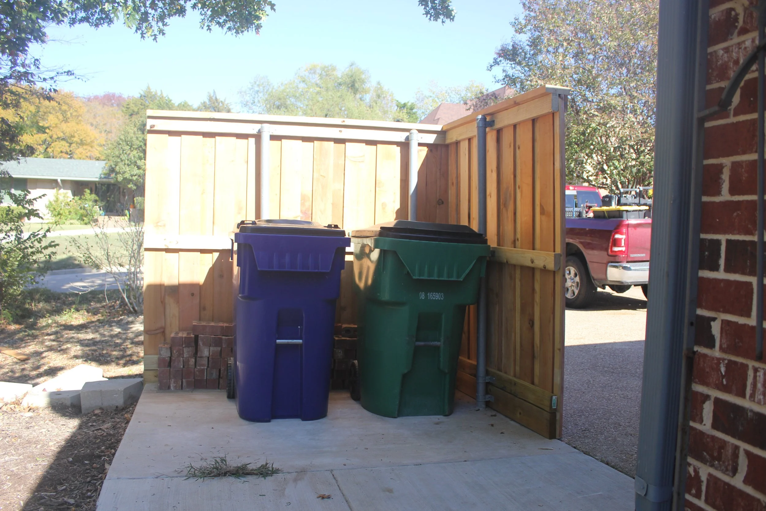 Two trash bins, one blue and one green, are placed next to a wooden fence on a concrete surface outdoors, with a red pickup truck and trees in the background.