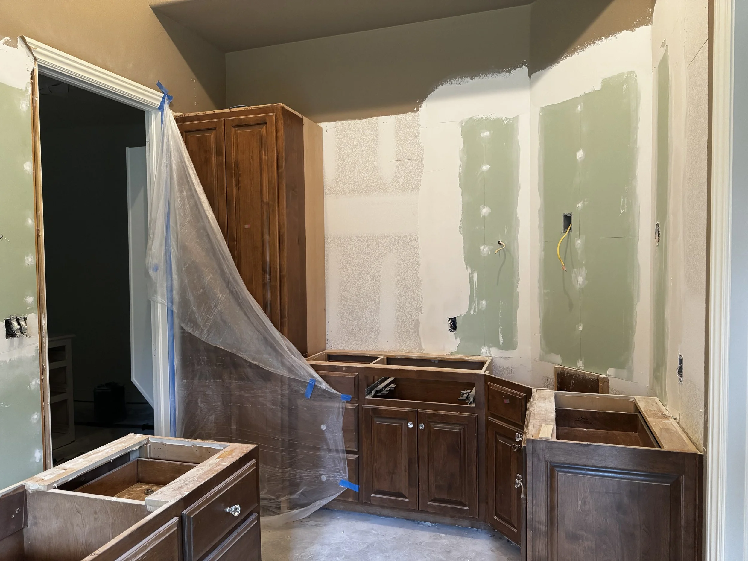 Kitchen under renovation with unfinished cabinets, exposed drywall, and wiring, partially covered with plastic sheeting.
