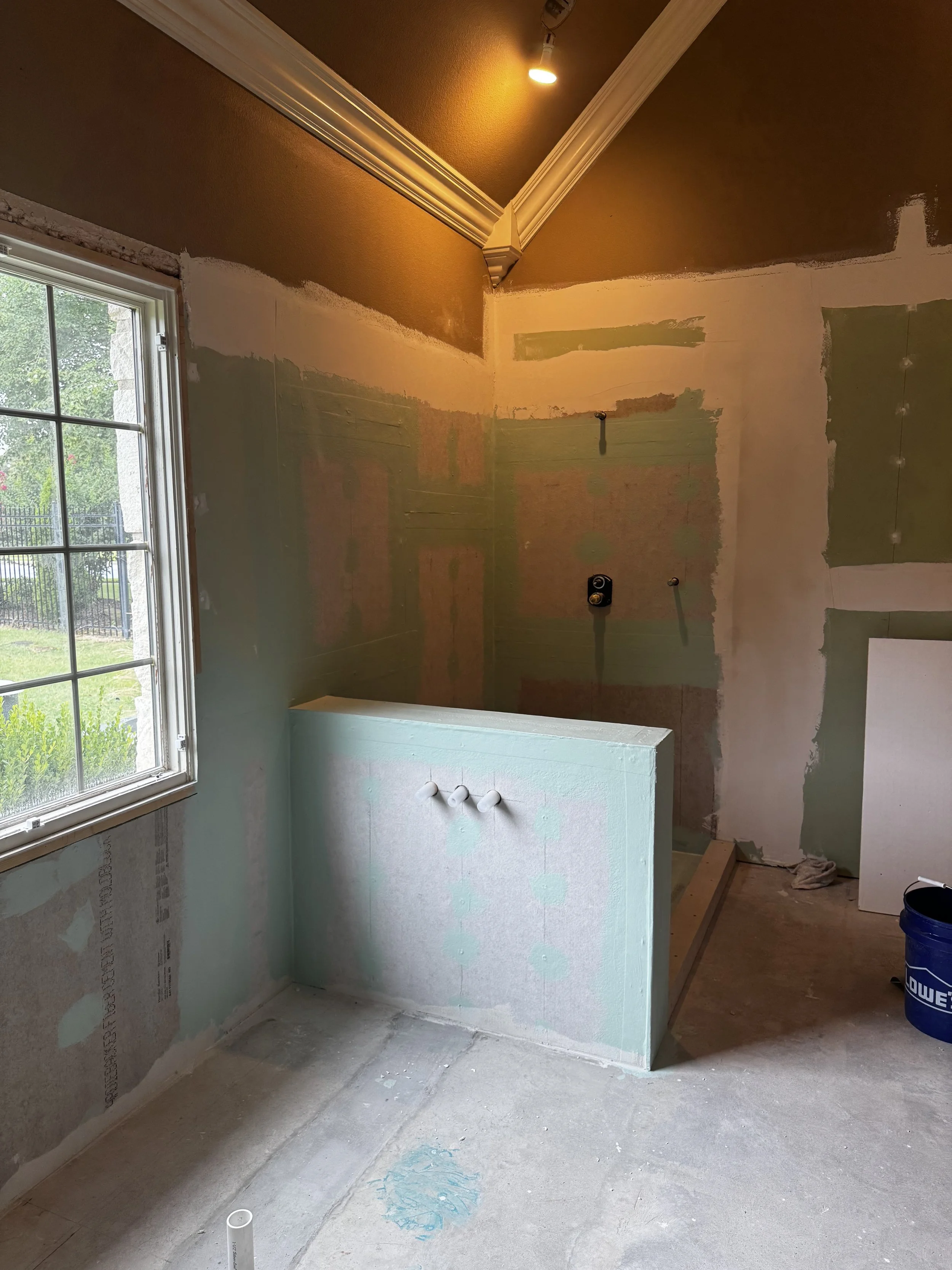 Bathroom under renovation with drywall partially installed, plumbing fixtures not yet connected, and construction materials on the floor. A window is on the left, and a ceiling light is turned on.
