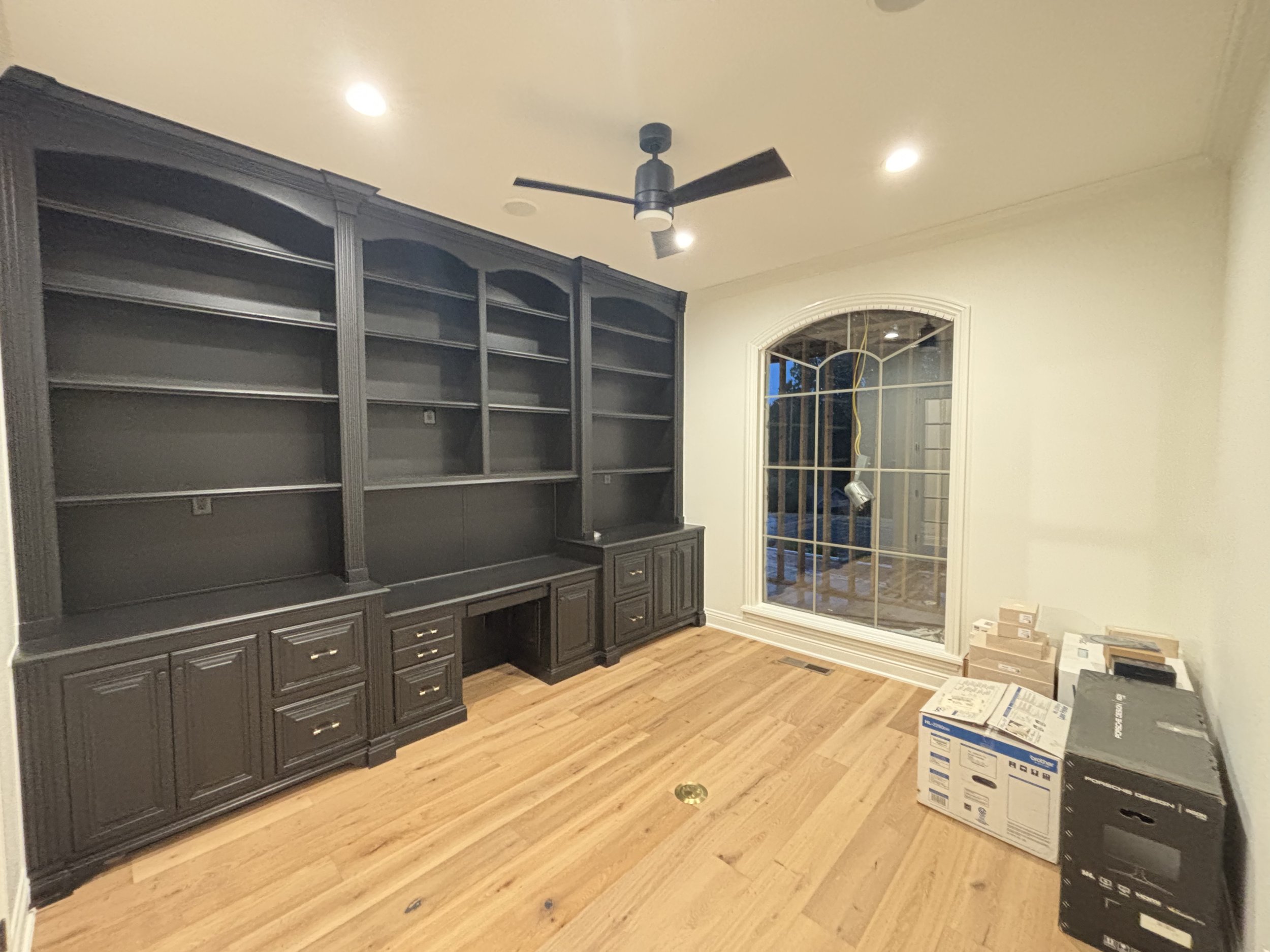 Empty room with dark built-in bookshelves, a ceiling fan, hardwood floors, and a large window with a grid design. There are boxes and a printer on the right side of the room.