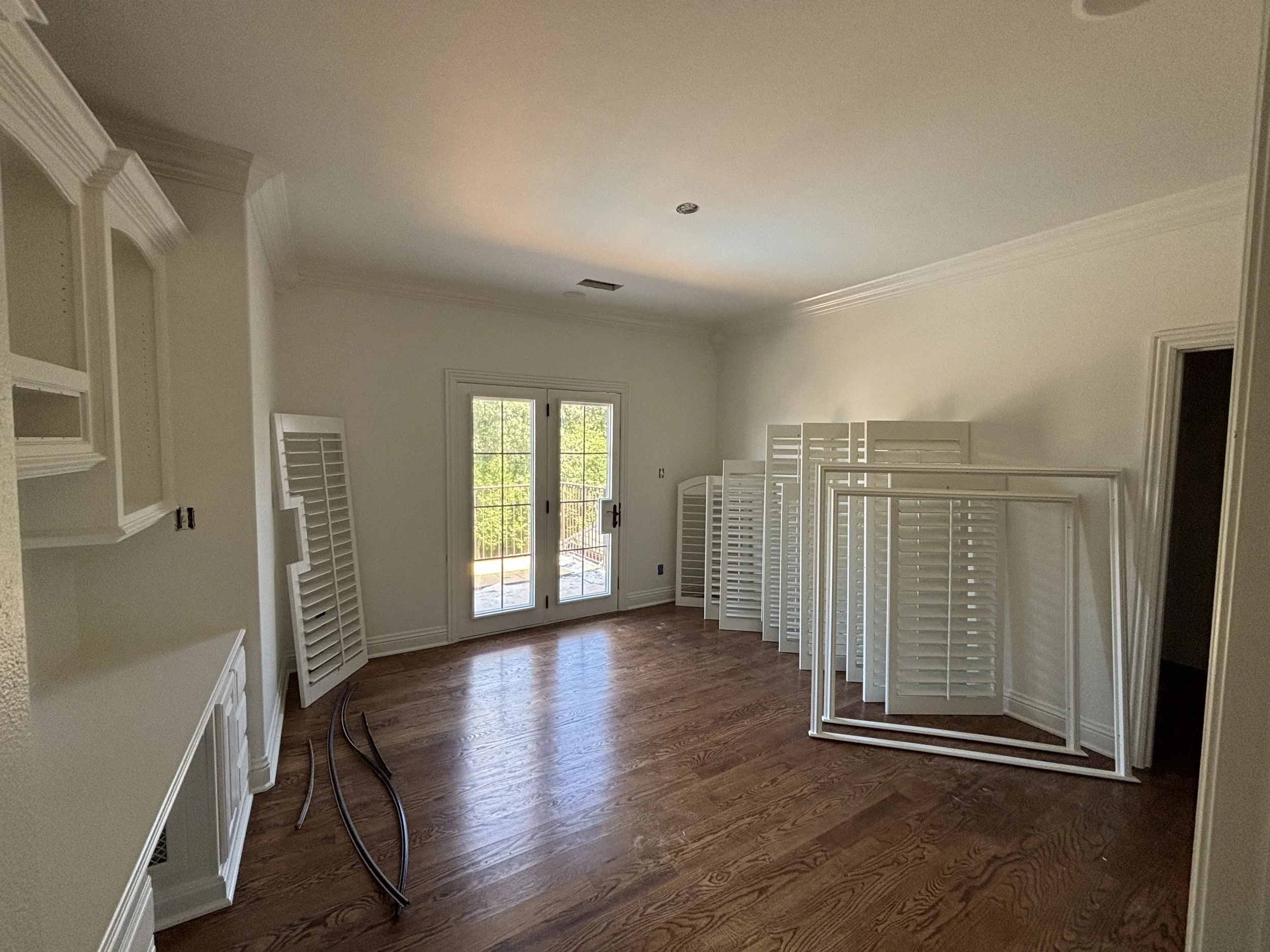 Empty room with hardwood floors, white walls, and French doors leading outside. Several white plantation shutters are leaning against the walls and on the floor, with some framing parts stacked next to the shutters.