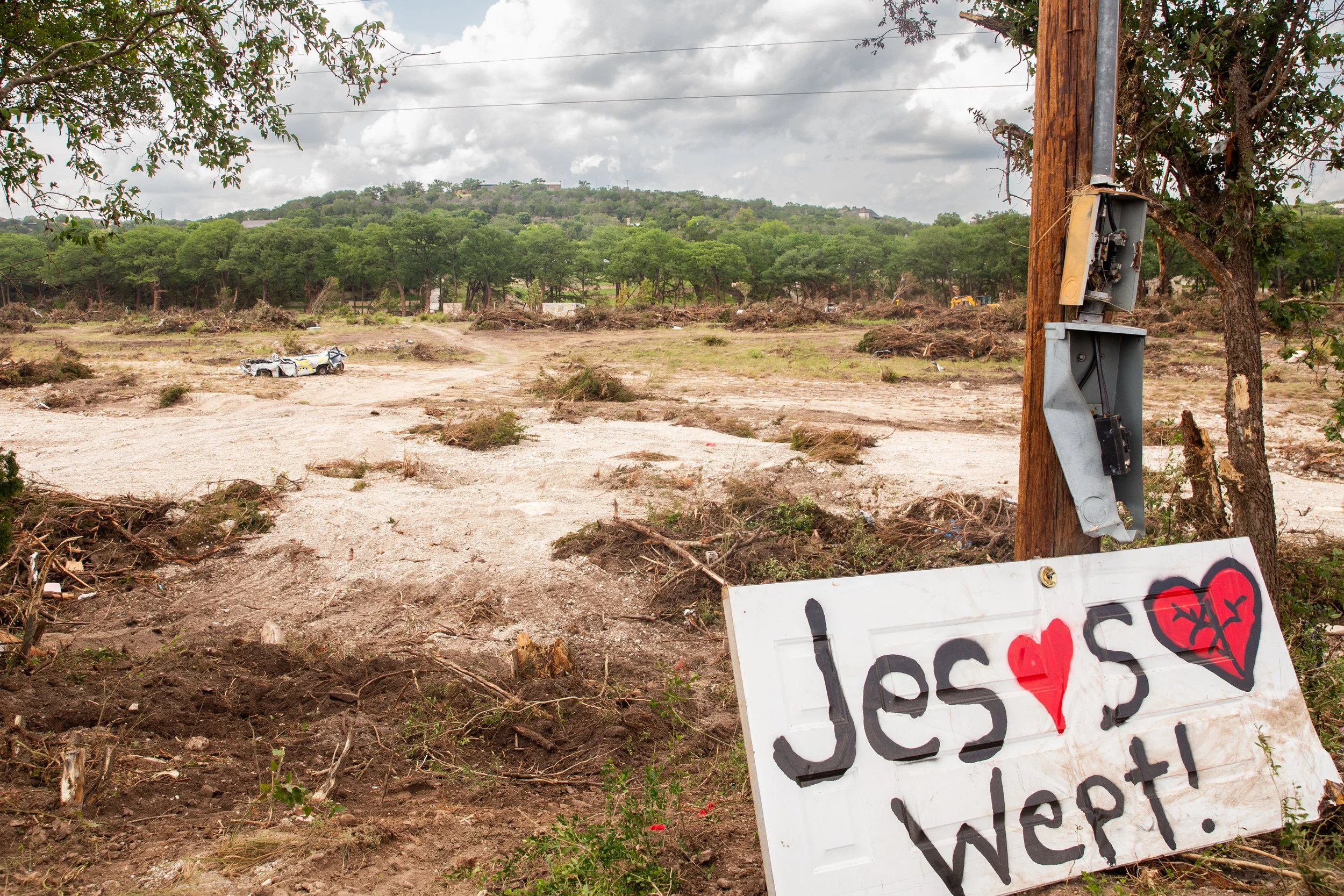 A damaged area of ground with tire tracks and debris, a fallen tree, and a wooden utility pole with an open electrical box. A white sign with colorful graffiti text 'Jesus ♥s Wep!' is leaning against the pole. In the background, there are green trees
